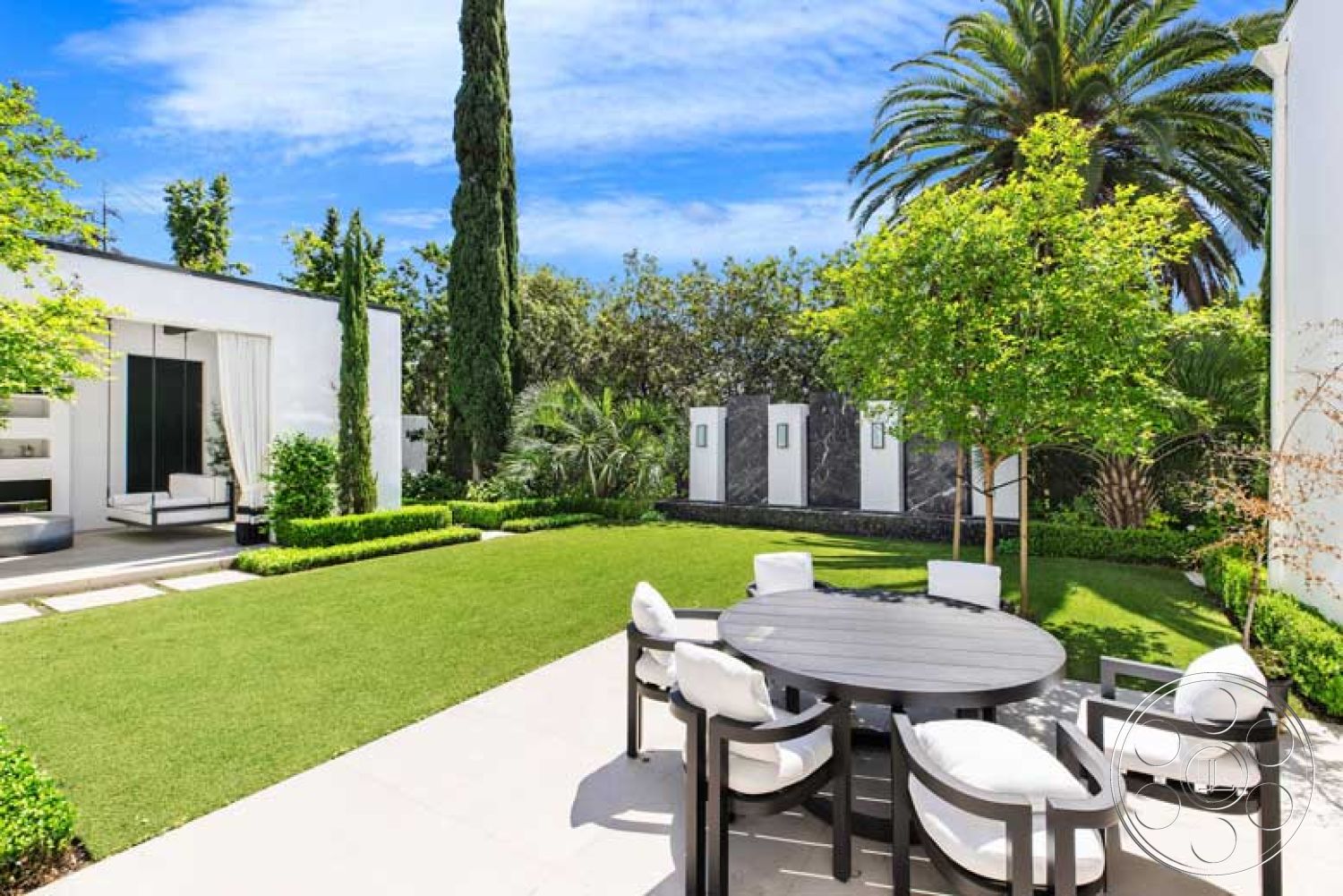 Modern 522 - exterior, floor-to-ceiling windows, lush green lawn, clear blue sky, cypress trees, rectangular dining table, tropical palm trees, ornamental shrubs, minimalist landscaping, modern outdoor seating, stone wall features, concrete patio floor, contemporary house facade, decorative garden wall