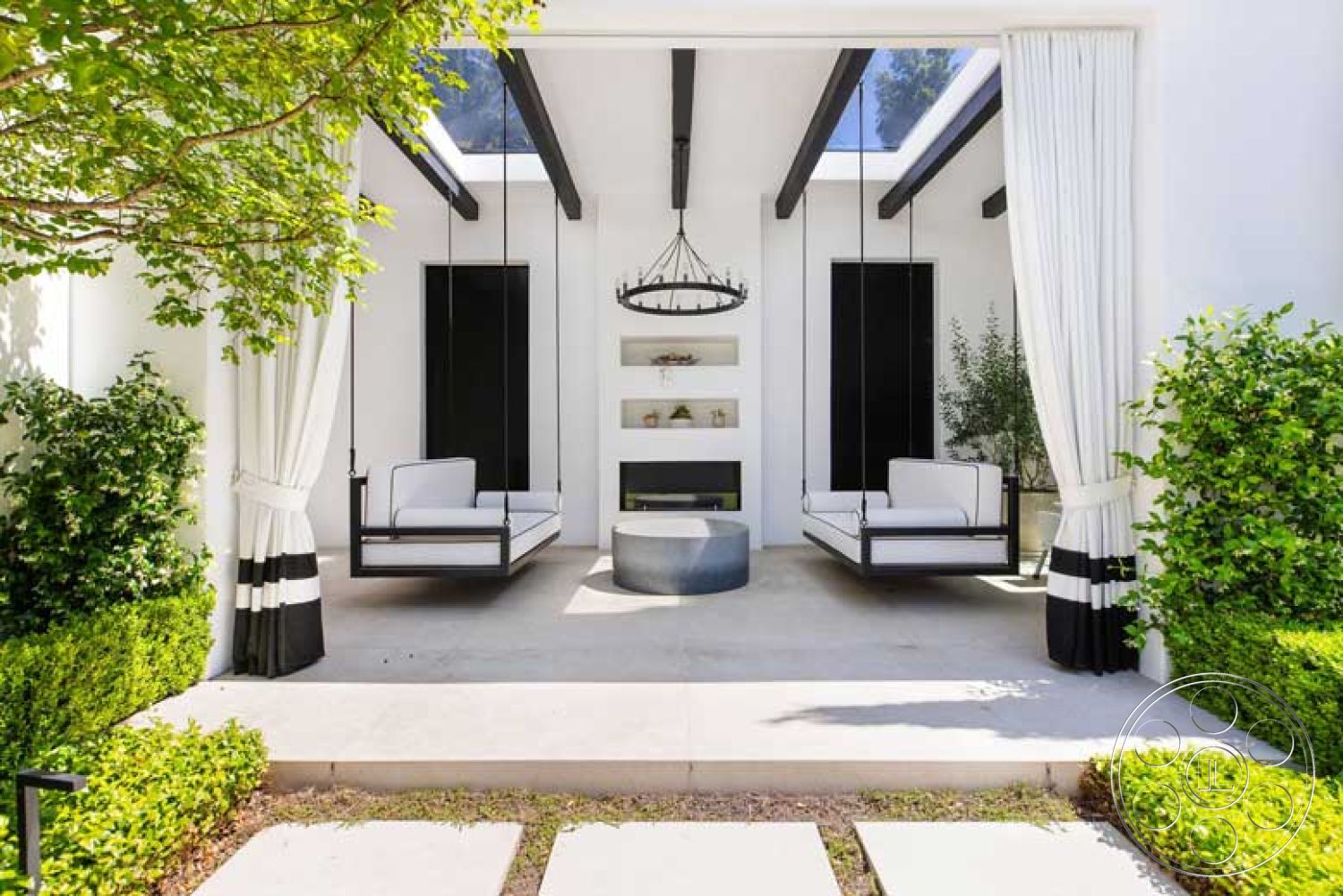 Modern 522 - exterior, green hedges, modern chandelier, black accent walls, exposed wood ceiling beams, concrete patio flooring, contemporary outdoor living area, hanging swing seating, minimalist fireplace feature wall, white drapery with black stripes, framed glass roof, paved stepping stones