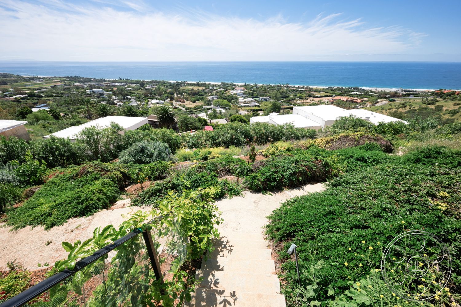 Modern 523 - residential neighborhood, hillside location, lush green landscaping, natural stone stepping stones, scenic coastal setting, outdoor garden path, overlooking ocean view, vibrant tropical plants, glass railing fence, open sky with wispy clouds, moderate slope terrain