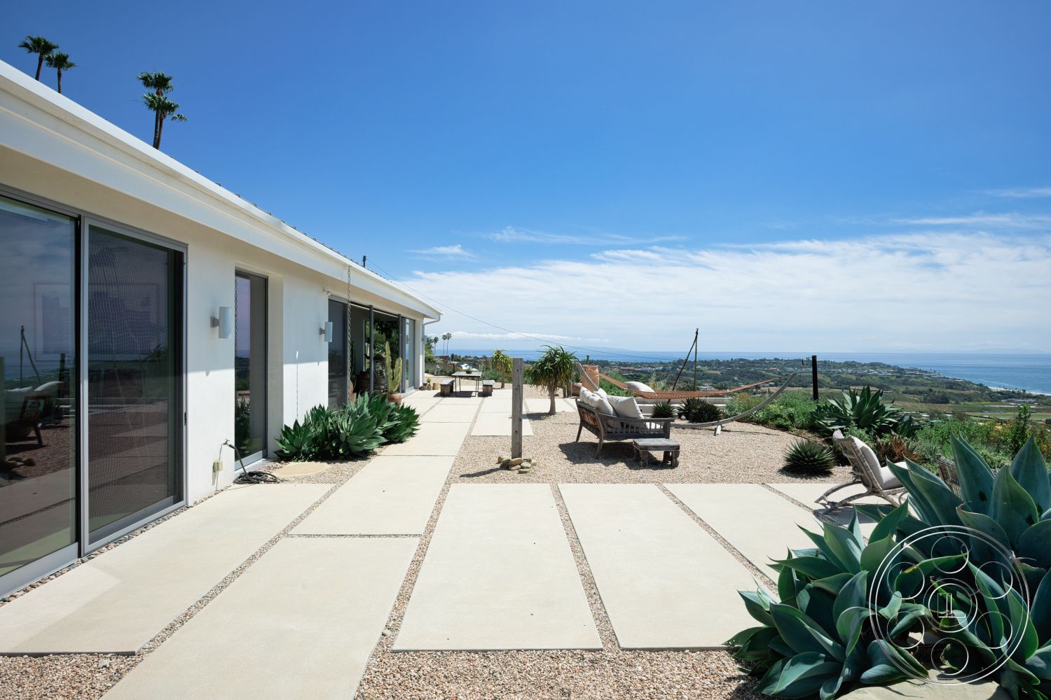 Modern 523 - exterior, large glass windows, smooth concrete surface, ocean view, concrete walkway, gravel landscape, lush green landscaping, white painted walls, modern minimalist architecture, sunny hillside location, desert home, low-profile roofline, patio sitting area, decorative agave plants
