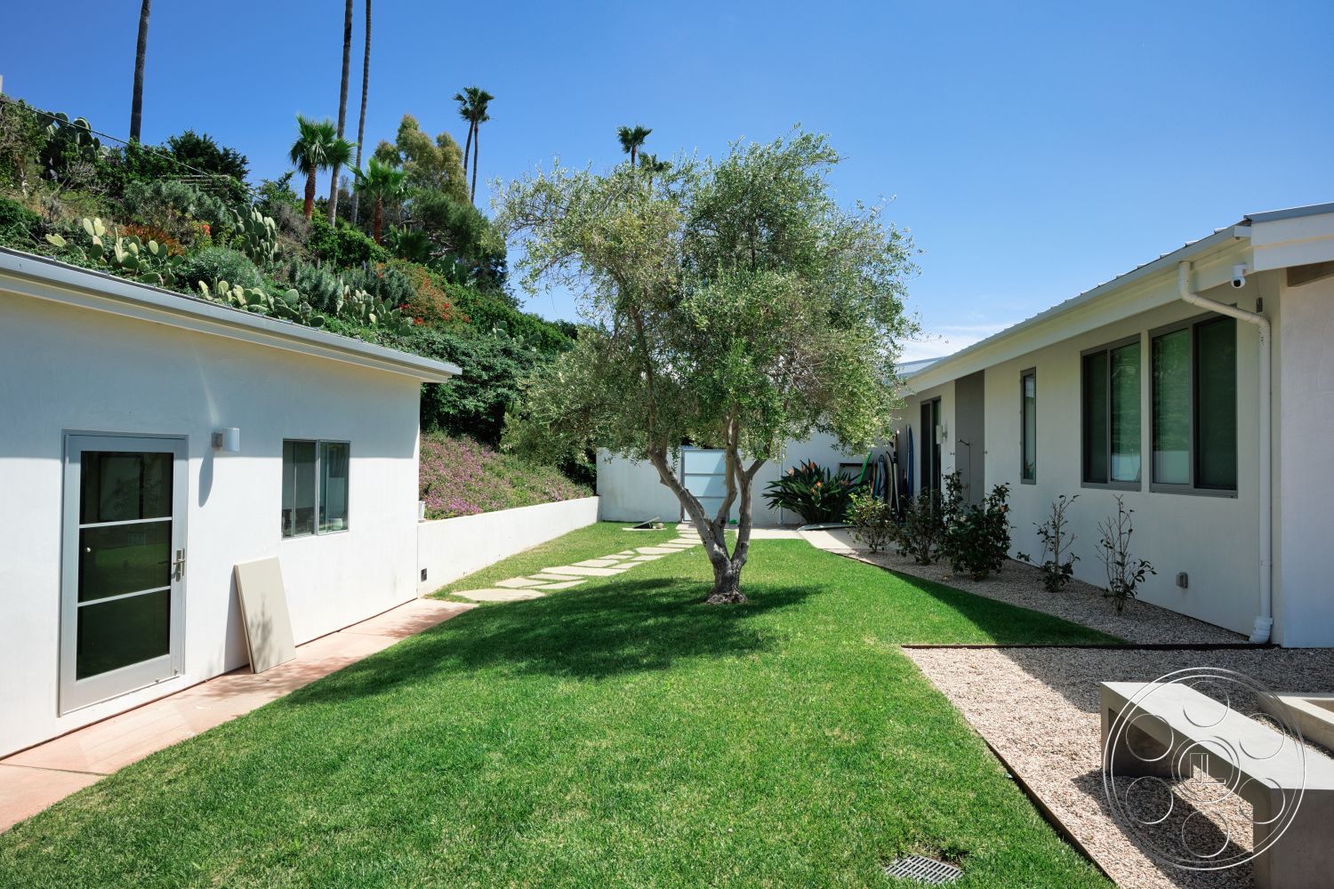 Modern 523 - large windows, exterior, white stucco walls, low-maintenance landscaping, green grass lawn, palm trees in background, flat roofing, modern home design, decorative gravel ground cover, sloped garden bed, concrete patio walkway, stone bench seating