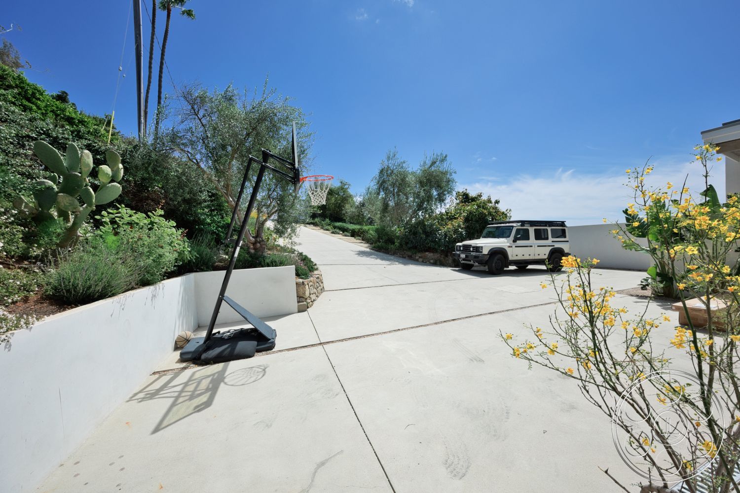 Modern 523 - exterior, textured concrete surface, landscaped garden area, open sky view, stamped concrete driveway, paved parking area, retaining wall with stone base, basketball hoop on portable stand, lush greenery with desert plants, gentle slope driveway