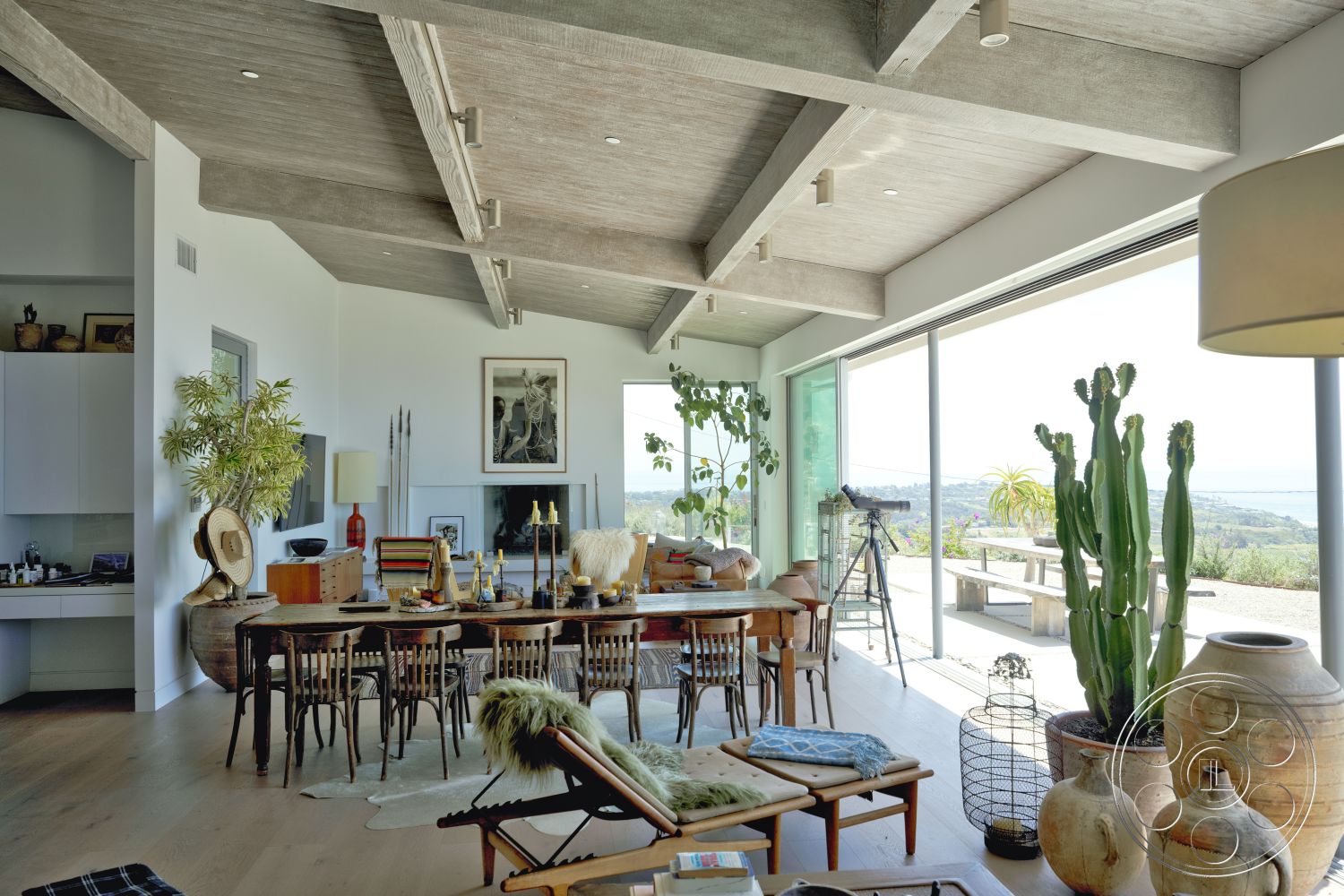 Modern 523 - textured area rug under dining table, modern pendant lighting fixtures, large floor-to-ceiling glass windows, exposed concrete ceiling with wooden beams, light hardwood flooring in living area, rectangular dining table with wooden chairs, minimalist white kitchen cabinets, natural stone countertop on kitchen island, decorative ceramic vases and sculptures, interior plants in decorative pots, open-concept living and dining space, contemporary art on feature wall, outdoor patio area visible through glass doors, urban hillside setting with ocean view