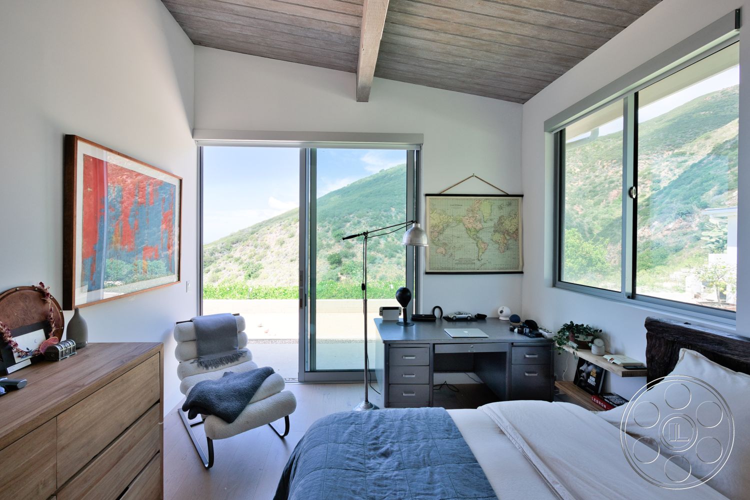 Modern 523 - large glass sliding doors, contemporary armchair, exposed wood ceiling beams, framed artwork on wall, textured white walls, light hardwood bedroom floors, modern desk with drawers, map wall art, mountain view from window