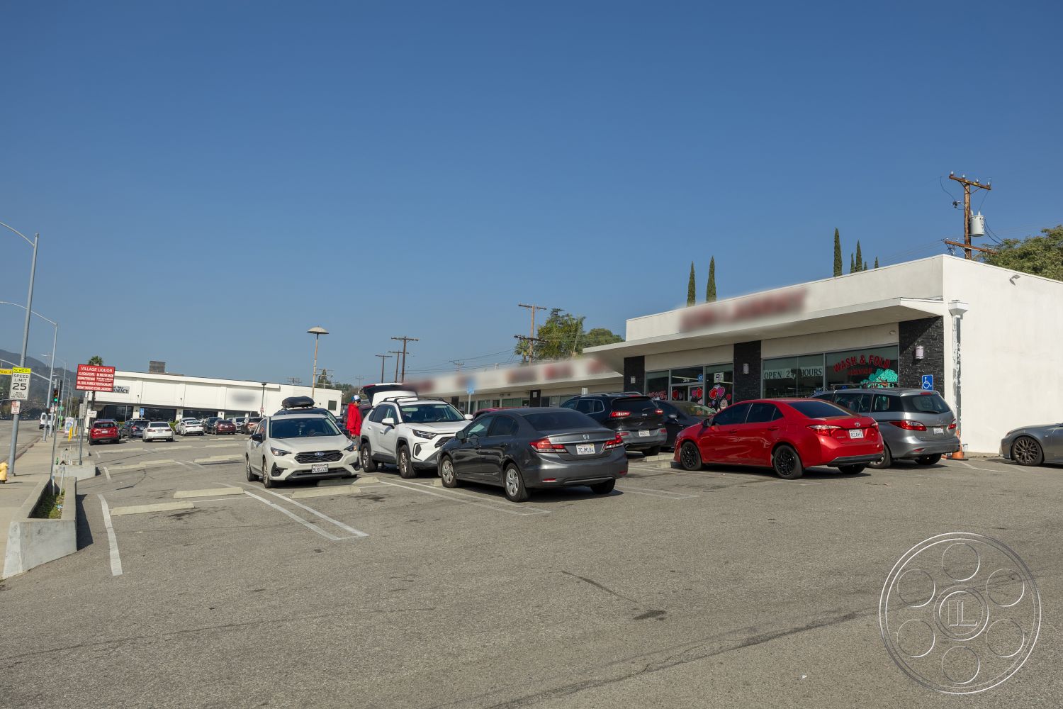 Laundromat 1 - paved driveway, landscaped planters, clear blue sky, concrete parking lot, glass storefront windows, urban commercial setting, exterior signage, flat roof commercial building, steel light poles, cypress trees landscaping, single-story retail space, parallel parking, open-air shopping area