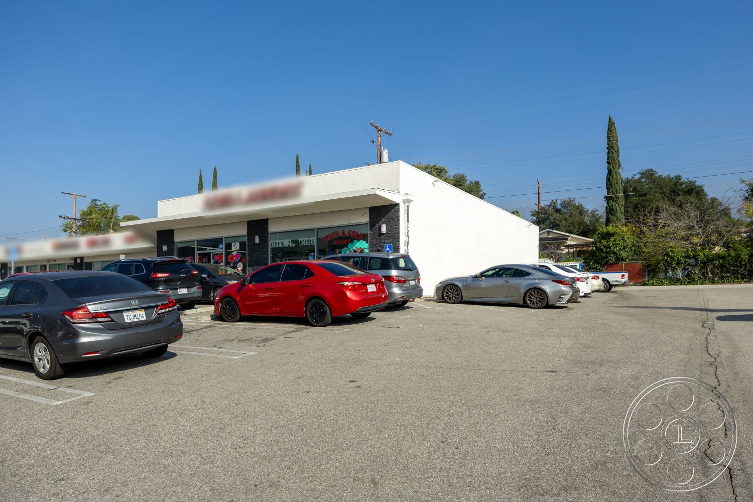 Laundromat 1 - exterior, flat roof, tall cypress trees, asphalt parking lot, concrete sidewalk, stucco building facade, large glass storefront windows, landscaped green spaces, strip mall architecture, gray asphalt with white parking lines