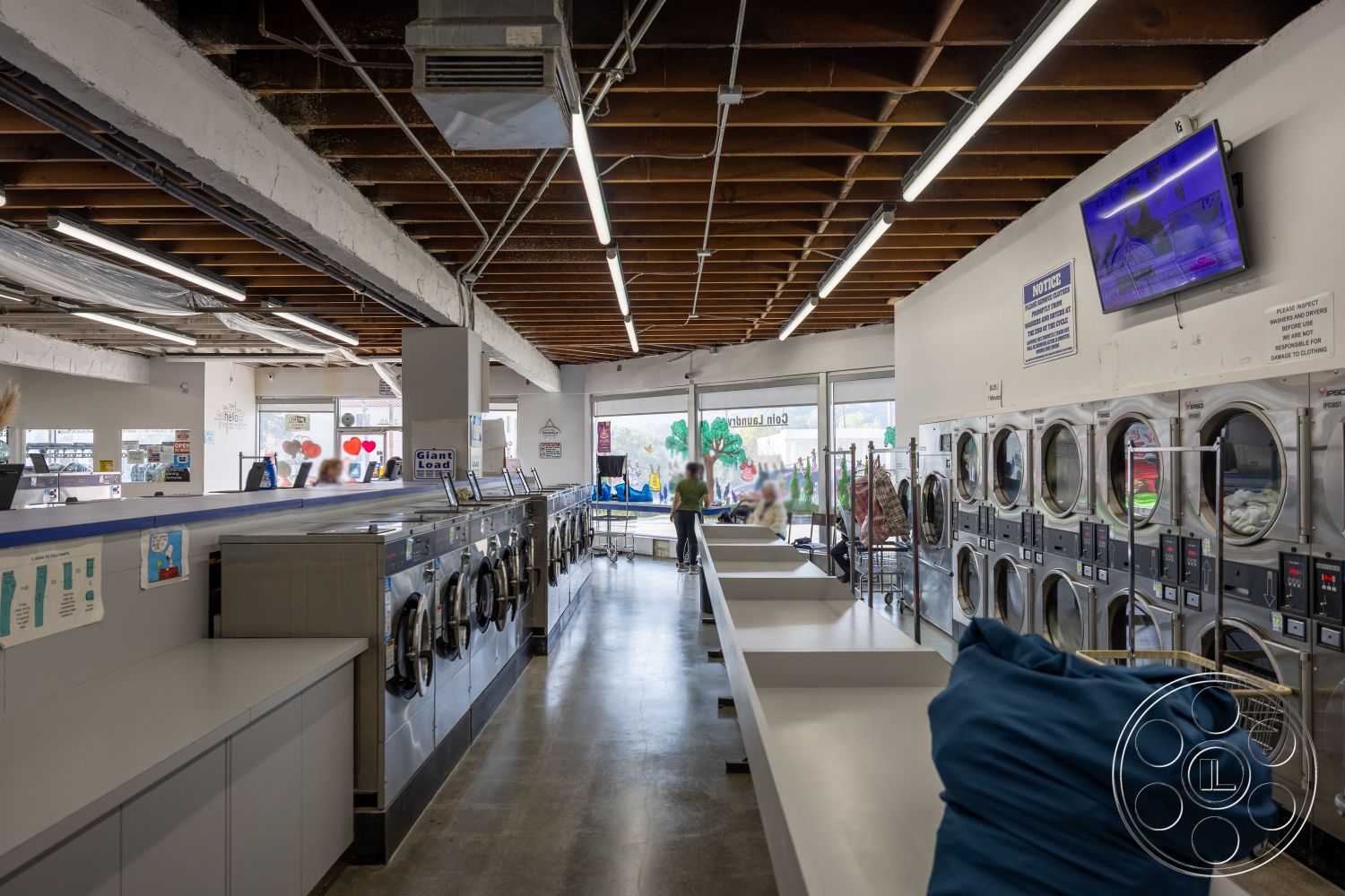 Laundromat 1 - open layout, concrete flooring, exposed wood ceiling beams, white painted walls, large windows with natural light, industrial style design, stainless steel countertops, fluorescent ceiling lighting, metal laundry machines, dryers with glass doors, customer seating area, laundry folding stations