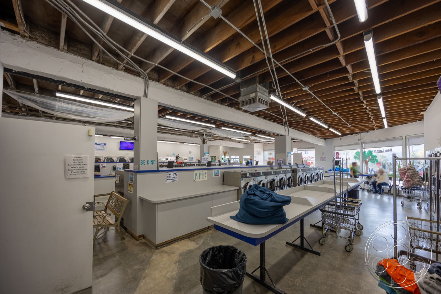 Laundromat 1 - concrete flooring, overhead fluorescent lighting, solid wood door, white painted walls, exposed wooden ceiling beams, glass storefront windows, informational signage, laundry folding tables, metal laundry carts, dryers and washing machines