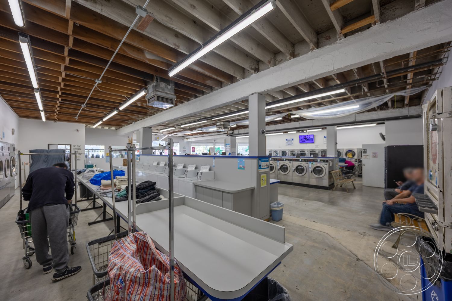 Laundromat 1 - open layout, concrete floor, seating area, laundry area, exposed wood ceiling beams, white painted walls, LED overhead lighting, metal laundry carts, commercial washing machines
