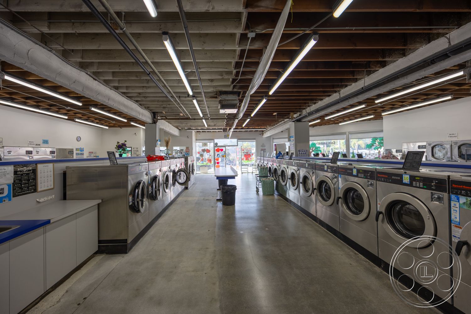 Laundromat 1 - white walls, concrete floor, modern lighting fixtures, exposed wood ceiling beams, glass storefront entrance, spacious interior layout, industrial-style design, stainless steel washing machines, laundry service counter, self-service laundromat