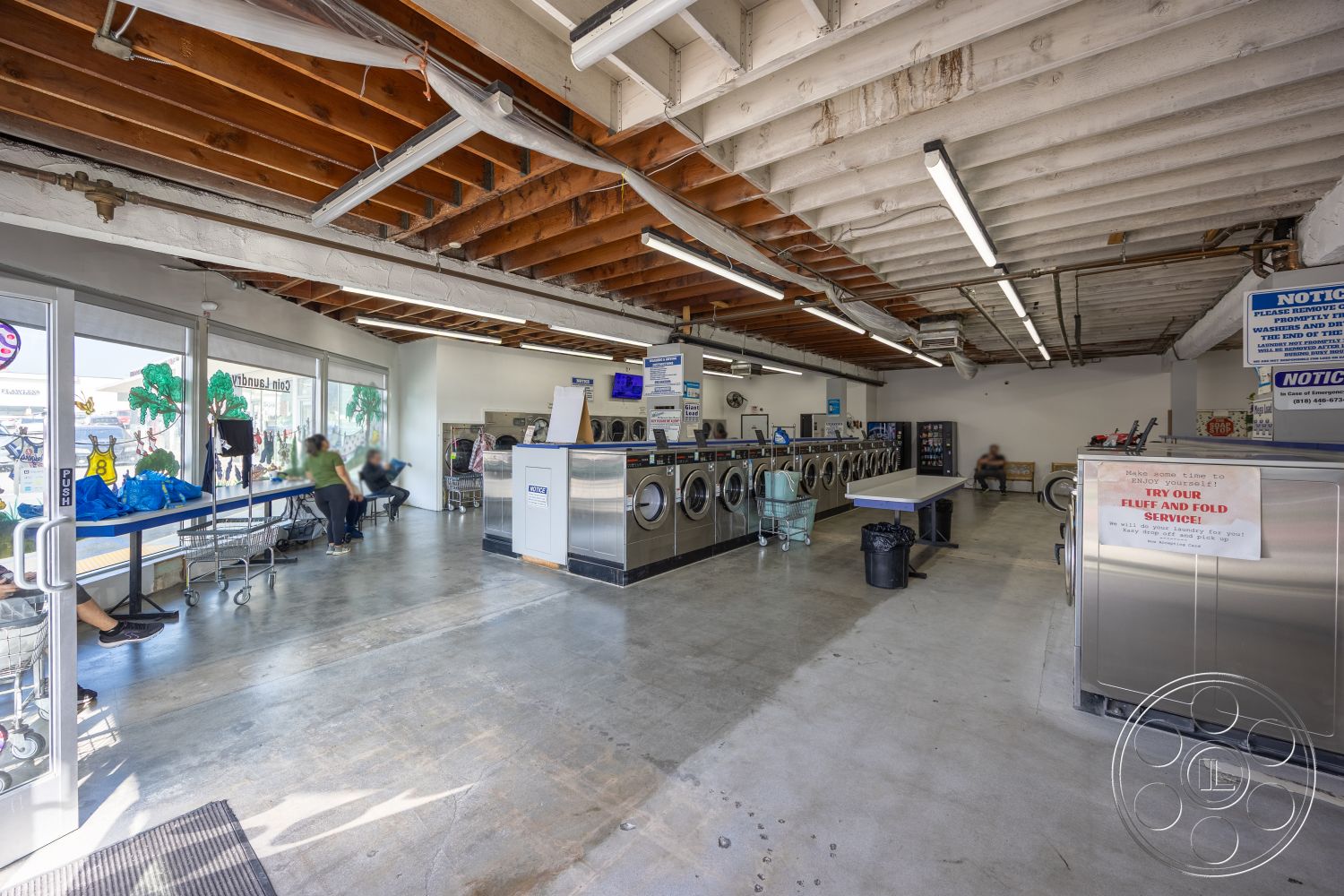 Laundromat 1 - open layout, polished concrete flooring, exposed wooden ceiling beams, glass entrance door, large windows providing natural light, fluorescent ceiling lighting, industrial laundry machines, customer seating area, laundry folding table, black plastic waste bin, machine-operated laundry area, signage for laundry services