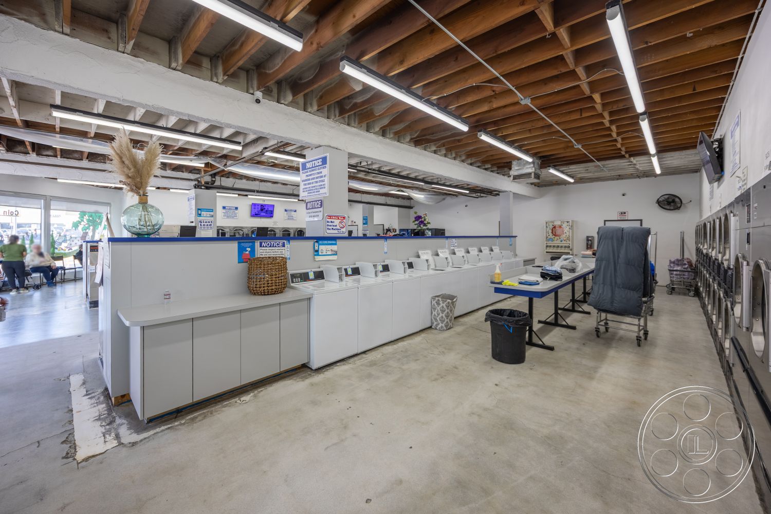 Laundromat 1 - concrete floor, exposed wood ceiling beams, open interior layout, fluorescent ceiling lights, blue accent walls, customer seating area, laundry counter cabinetry, white laundry machines, reception desk space, industrial laundry setting