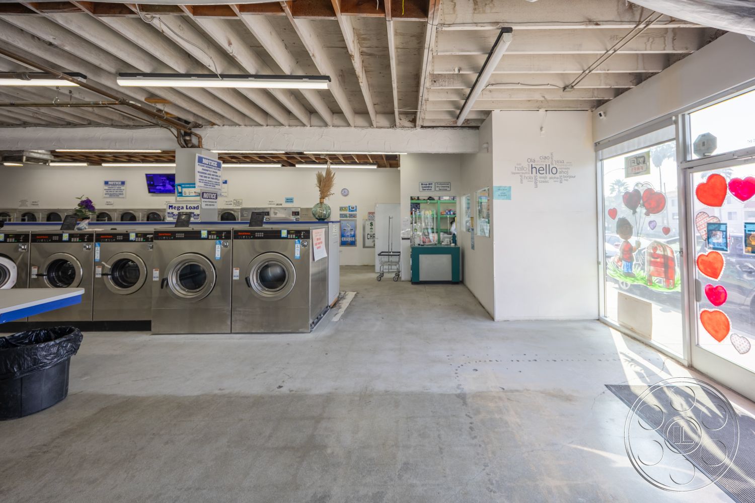 Laundromat 1 - open layout, concrete floor, industrial lighting fixtures, white painted walls, exposed wooden ceiling beams, glass entrance door, stainless steel washing machines, customer seating area, laundry service area