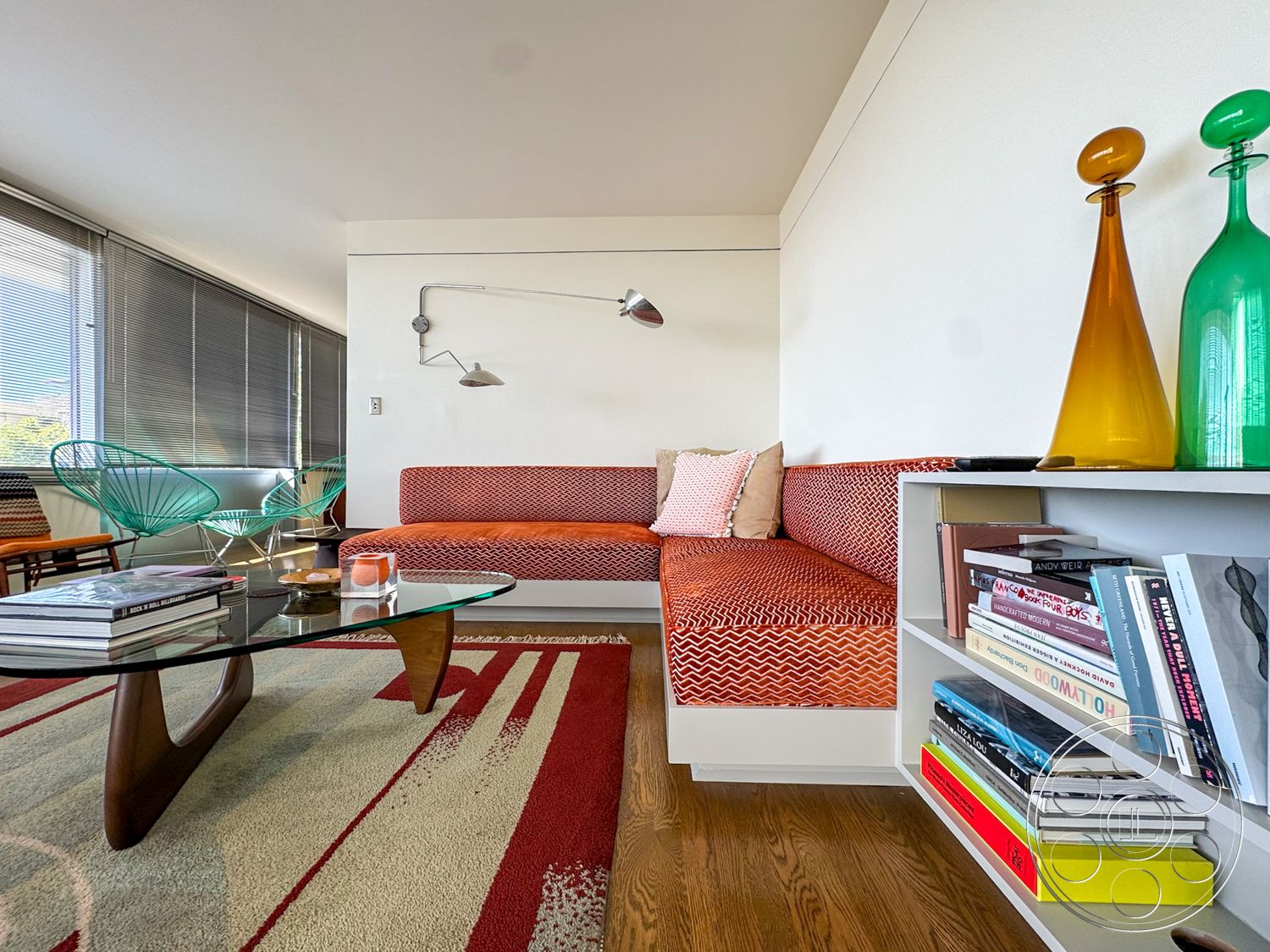 The Kun House - wood flooring, neutral wall colors, textured area rug, low-profile sofa, glass coffee table, modern design elements, white ceiling, built-in bookshelf, mid-century modern furniture, curved lounge chairs, large windows with blinds, orange patterned upholstery, acrylic table base, decorative glass vases, metal wall sconce lighting