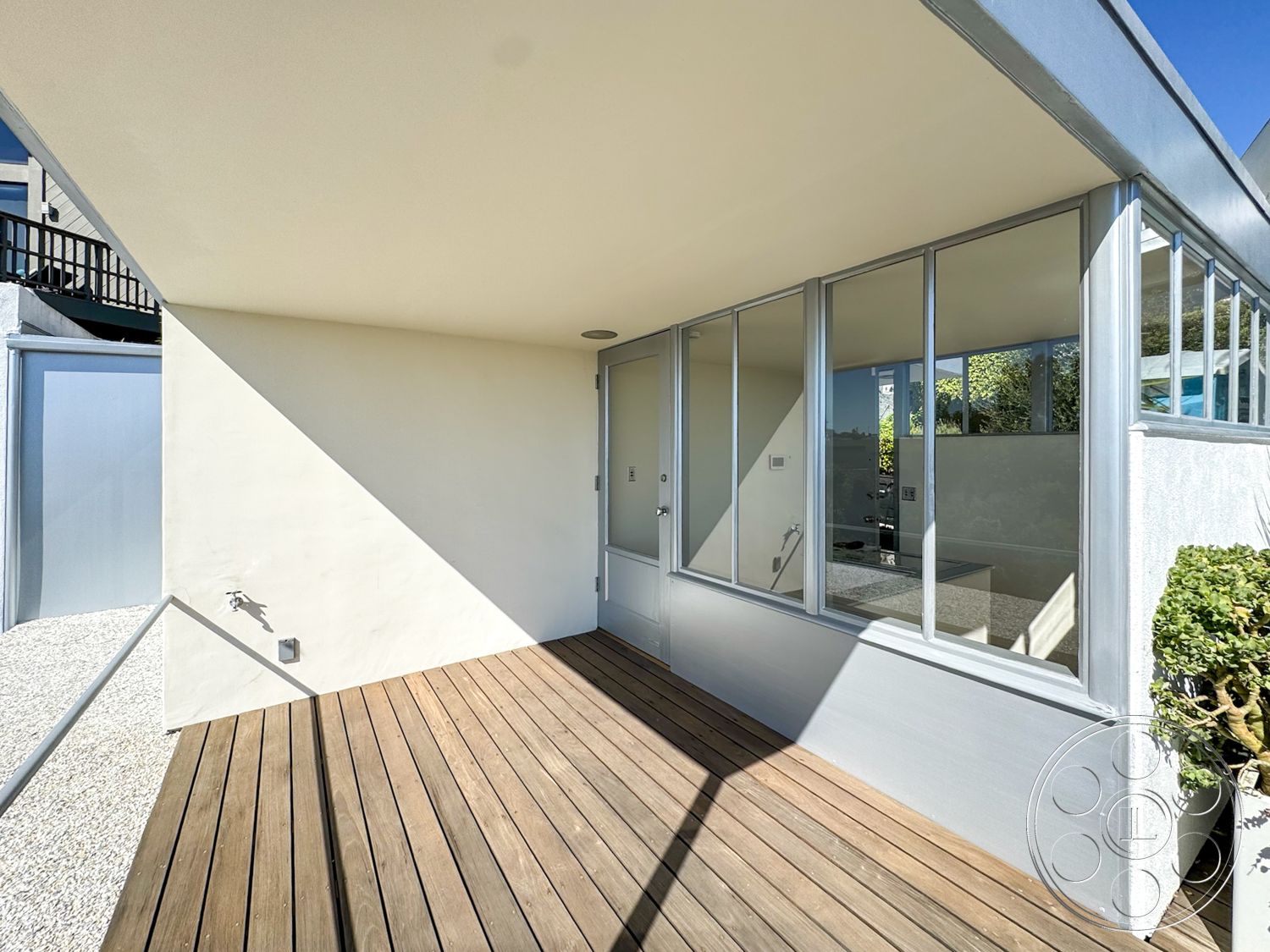 The Kun House - exterior, large glass windows, outdoor seating area, metal railing, landscaped greenery, modern architectural style, wooden deck flooring, stucco wall finish, concrete boundary wall, angular roofline