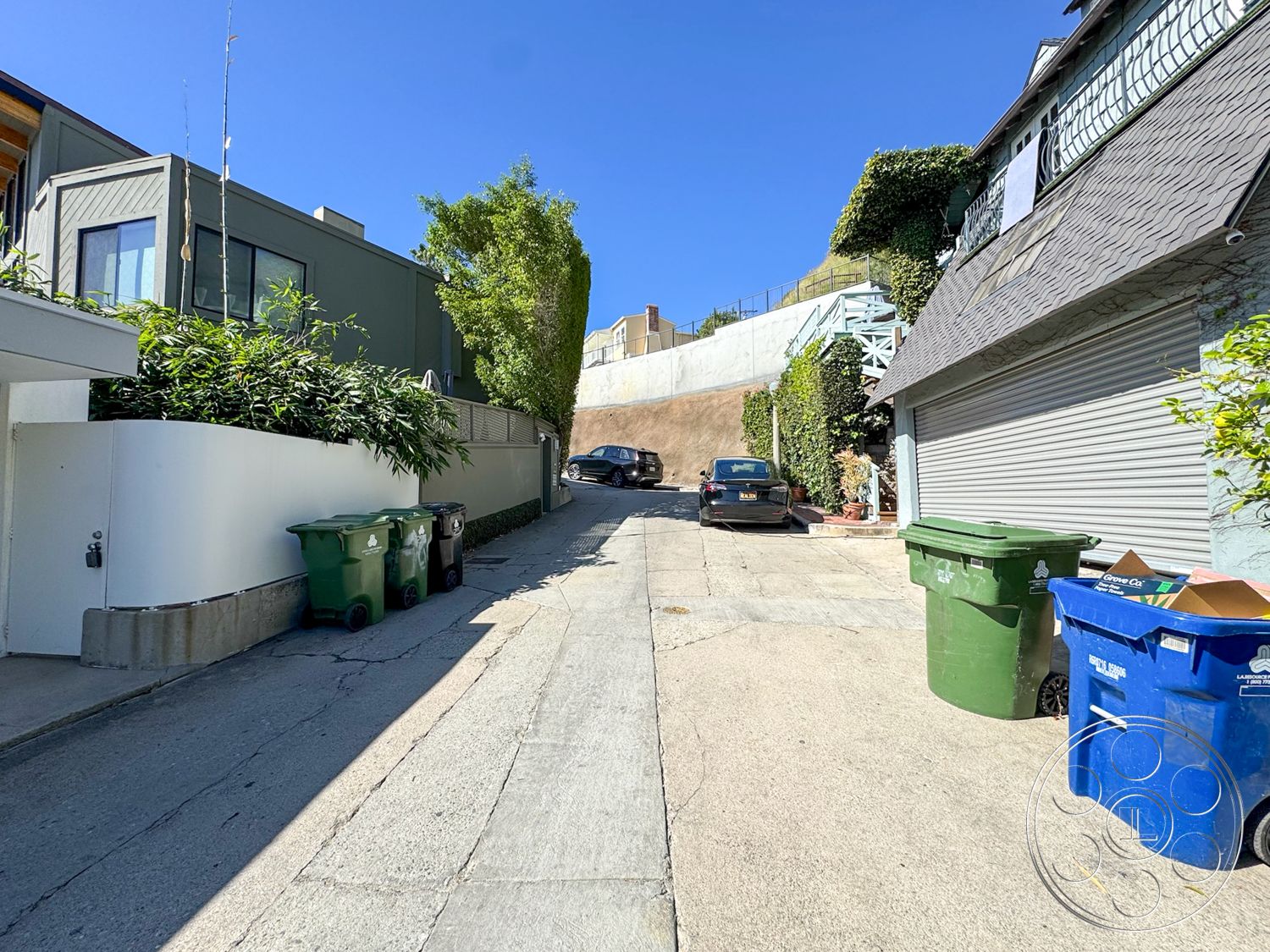 The Kun House - exterior, sloped terrain, concrete driveway, green trash bins, urban alley setting, asphalt roadway, landscape vegetation, modern architecture style, metal garage door, exposed hillside, brick privacy wall, blue recycling bin