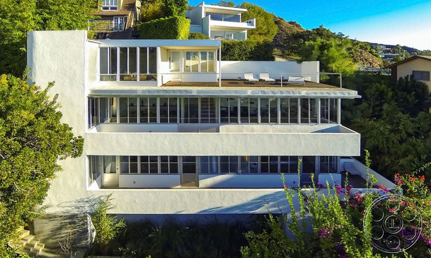 The Kun House - exterior, white stucco walls, floor-to-ceiling windows, modern architectural style, surrounded by greenery, hillside location, rectangular building shape, horizontal balcony railings, flat roof deck, three-story contemporary home, open terrace with loungers, sunny exterior facade