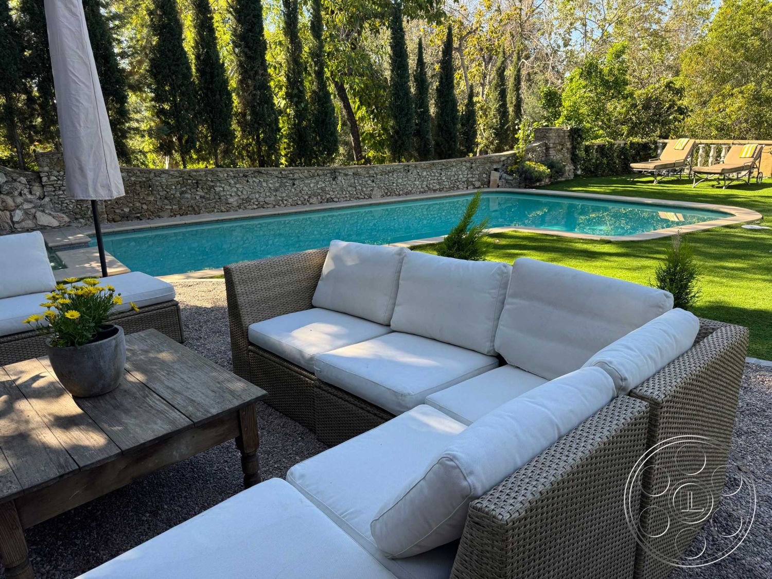 Medit 156 - exterior, landscaped garden, wood coffee table, rectangular swimming pool, lawn area, tall cypress trees, sun umbrella, gravel patio flooring, woven outdoor sofa, stone pool border, stone wall garden perimeter, fabric cushions, potted flowering plant, sun lounger deck chairs