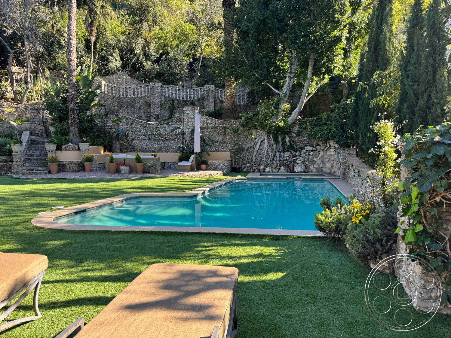 Medit 156 - exterior, lush greenery, outdoor seating area, tropical landscaping, residential backyard, artificial turf lawn, rectangular pool, wooden lounge chairs, natural stone retaining wall, landscaped garden area, concrete pool border, potted plants with flowers, sunny backyard setting, curved stone steps