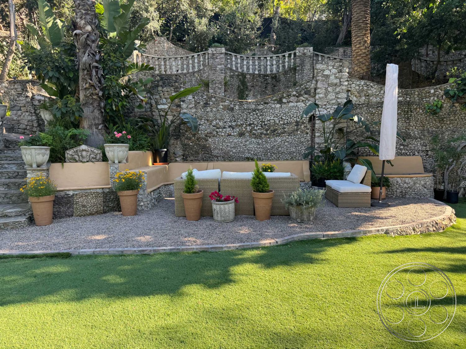 Medit 156 - exterior, tropical plants, terracotta planters, lush tropical landscaping, outdoor relaxation area, gravel patio flooring, green lawn area, potted flower arrangements, rough stone wall, light-colored seating cushions, curved patio seating structure, smooth stone steps, parasol umbrella, vertical stone garden wall, decorative garden railing