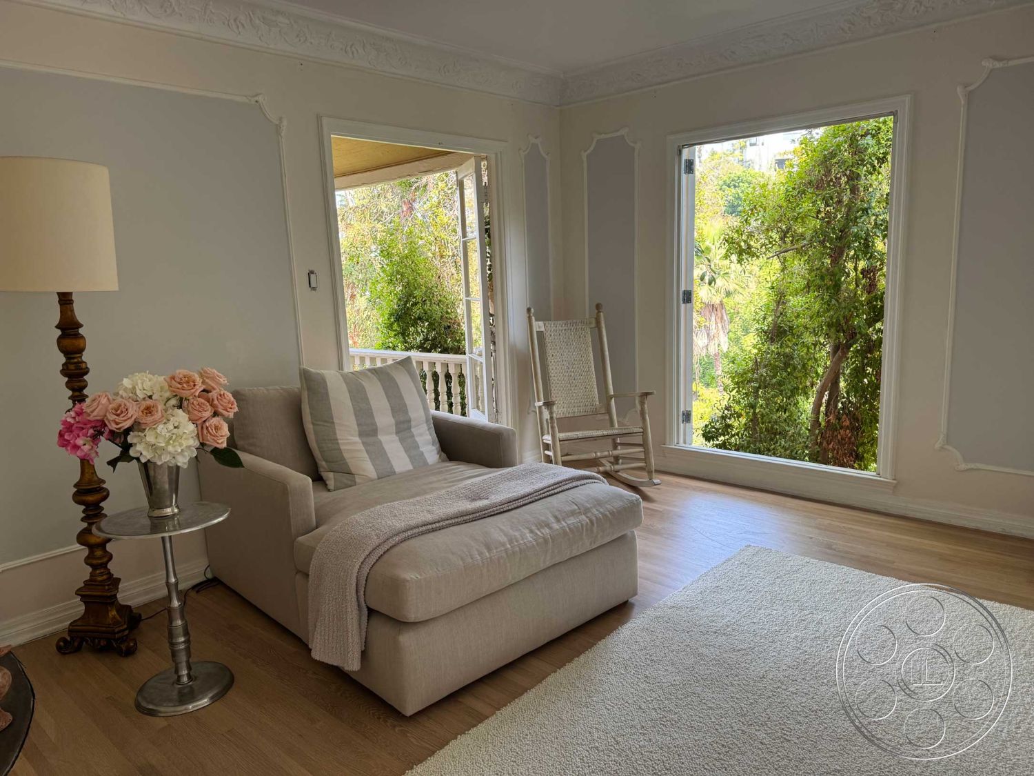 Medit 156 - soft natural lighting, light wood floor, white painted walls, traditional rocking chair, intricate crown molding, beige upholstered chaise lounge, striped throw pillow, textured beige throw blanket, white decorative rug, glass window with nature view, silver metal side table, elegant flower arrangement