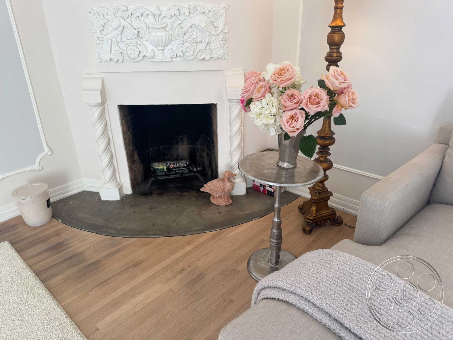 Medit 156 - white plaster walls, hardwood living room floors, exposed brick fireplace surround, ornate wall molding, curved concrete fireplace hearth, decorative metal side table, modern beige sofa, vase with pink roses, light wood lamp, textured gray throw blanket