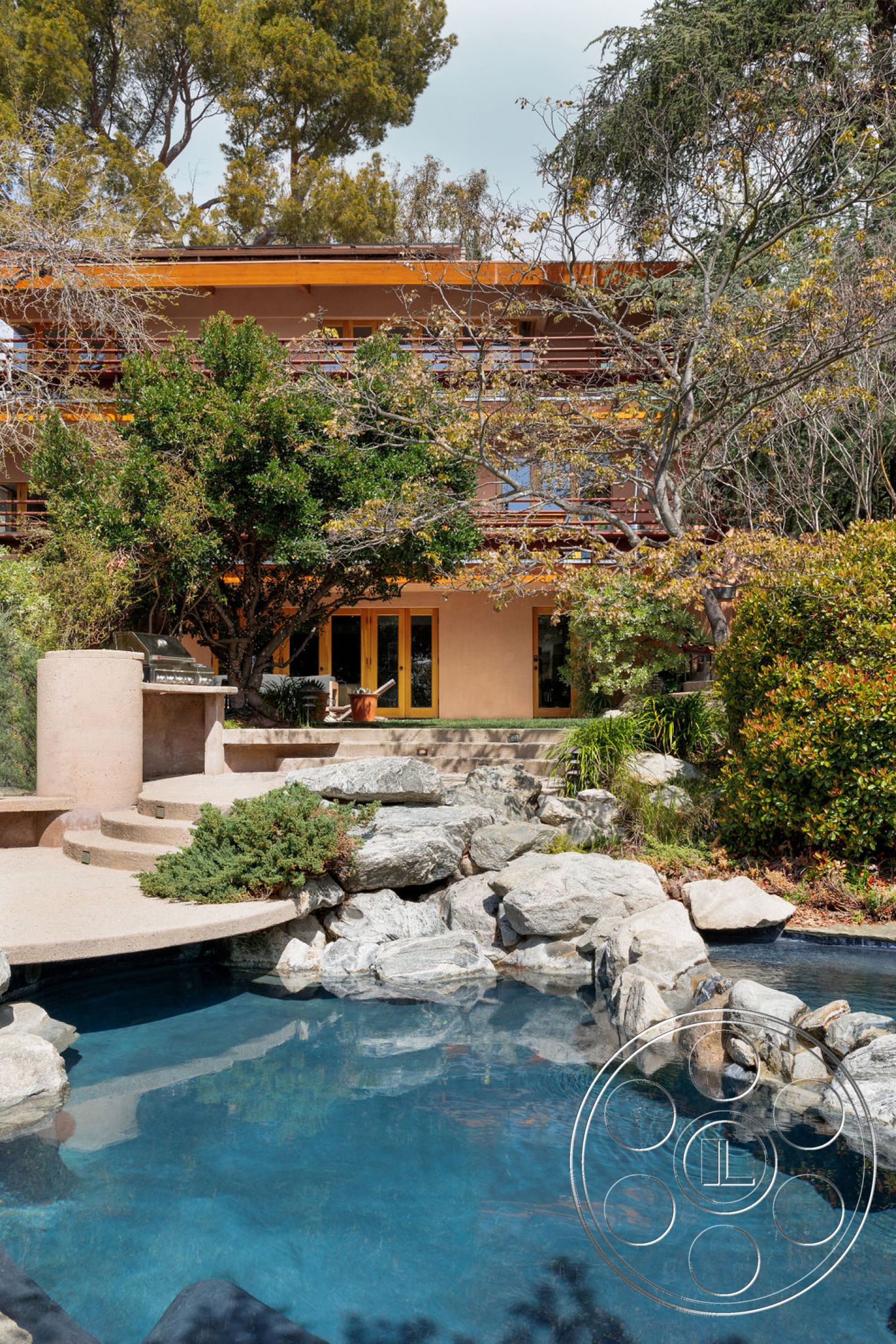 Mid-Century Exotic - exterior, glass sliding doors, wooden deck, rectangular swimming pool, tree canopy, outdoor kitchen, tan stucco walls, landscaped garden area, concrete pool surround, natural rock landscaping, stone fountain feature, yellow window frames, multi-level deck