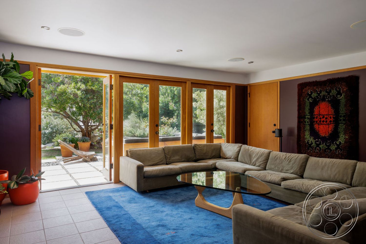 Mid-Century Exotic - textured wall art, open floor plan, glass coffee table, green indoor plants, natural light from large windows, wooden door frames, light-colored ceramic tile floor, beige upholstered sectional sofa, wood-framed sliding glass doors, hidden ceiling speakers, blue area rug, landscaped outdoor garden view