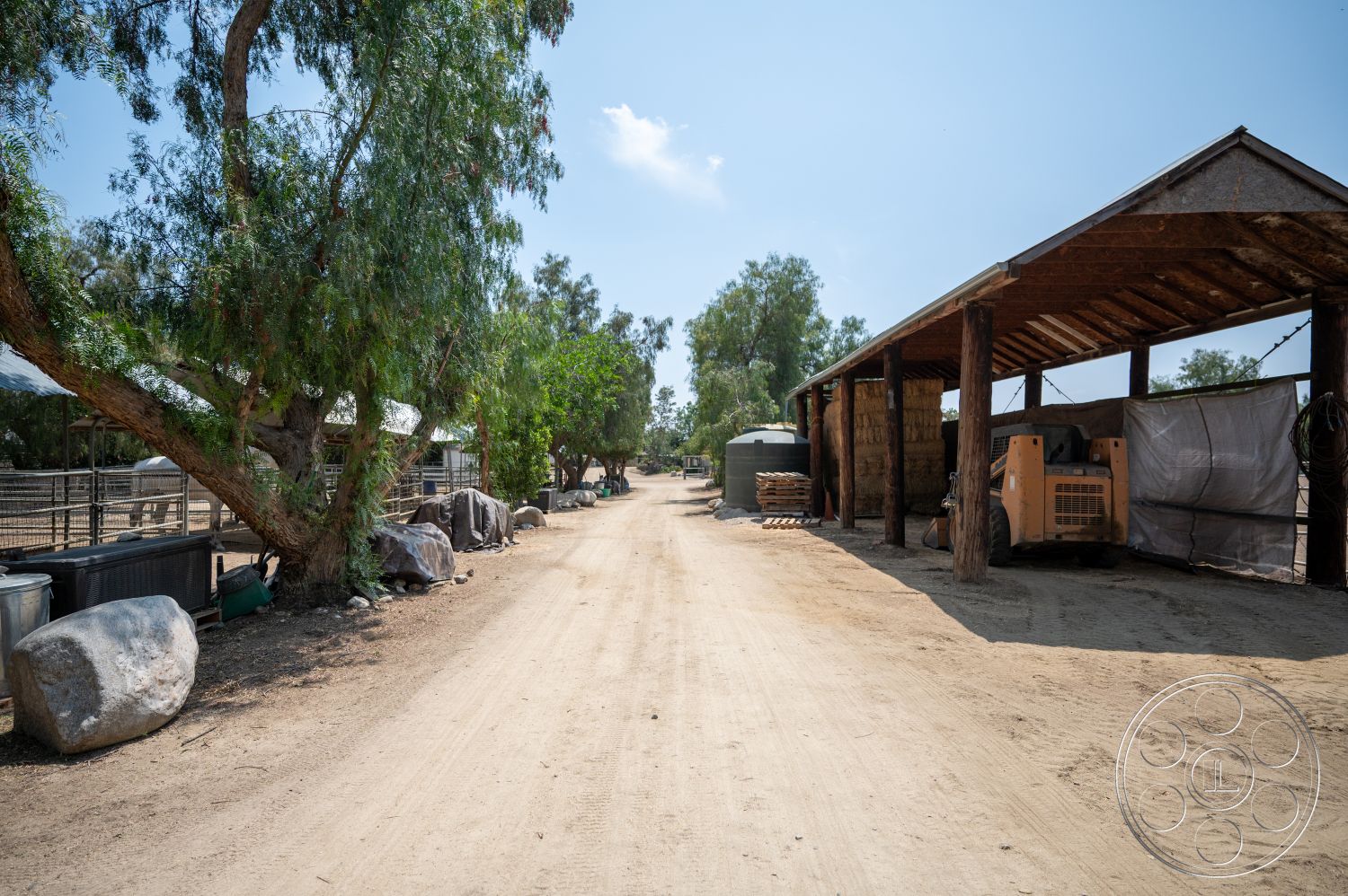 Ranch 6 - exterior, clear blue sky, dirt driveway, rural landscape, lush green trees, metal storage tanks, weathered wooden beams, farm environment, hay bales, wooden shelter roof, round stone boulders, open ranch setting