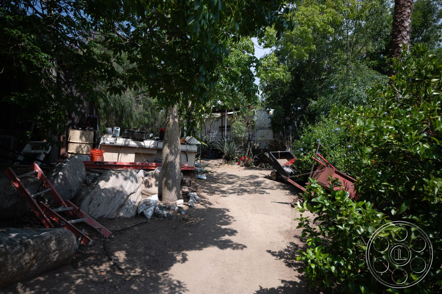 Ranch 6 - exterior, landscaped garden, potted plants, tree canopy, wooden ladder, gravel pathway, green shrubs, dirt ground, decorative flowers, concrete block wall, natural stone boulders, rusted metal wheelbarrow, building structure