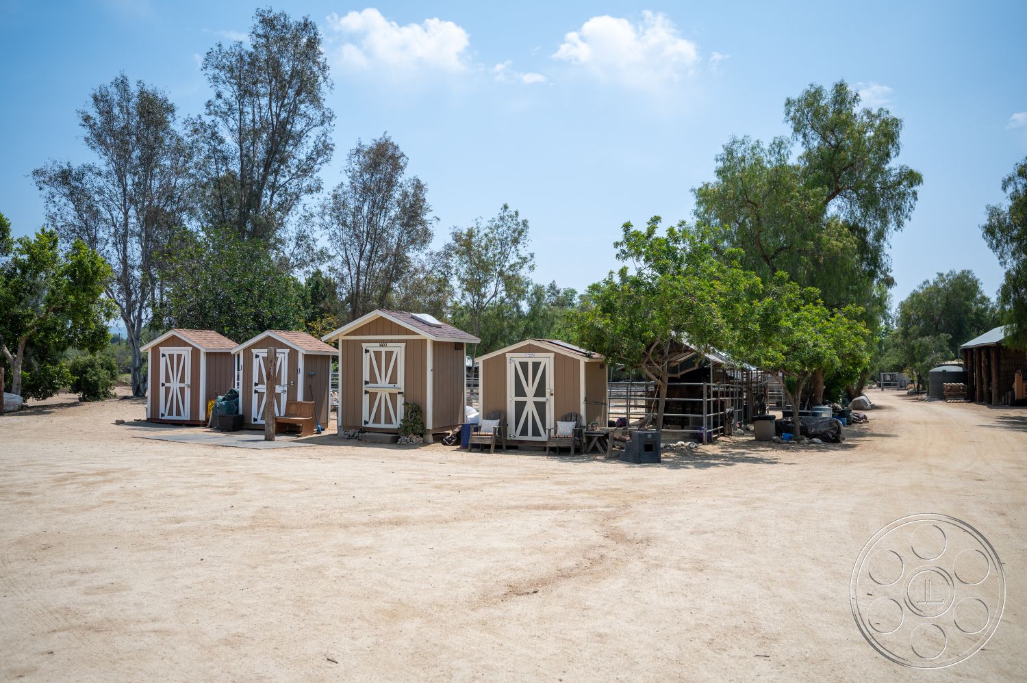Ranch 6 - exterior, surrounding trees, dirt parking area, double door entry, rural farmhouse setting, gravel access path, wooden storage sheds, light tan wooden siding, brown wooden roofs
