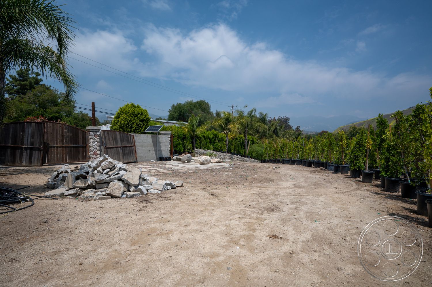Ranch 6 - exterior, wooden fence, gravel driveway, stone retaining wall, potted shrubs, dirt landscaping, sunny suburban yard, porous soil, construction materials pile