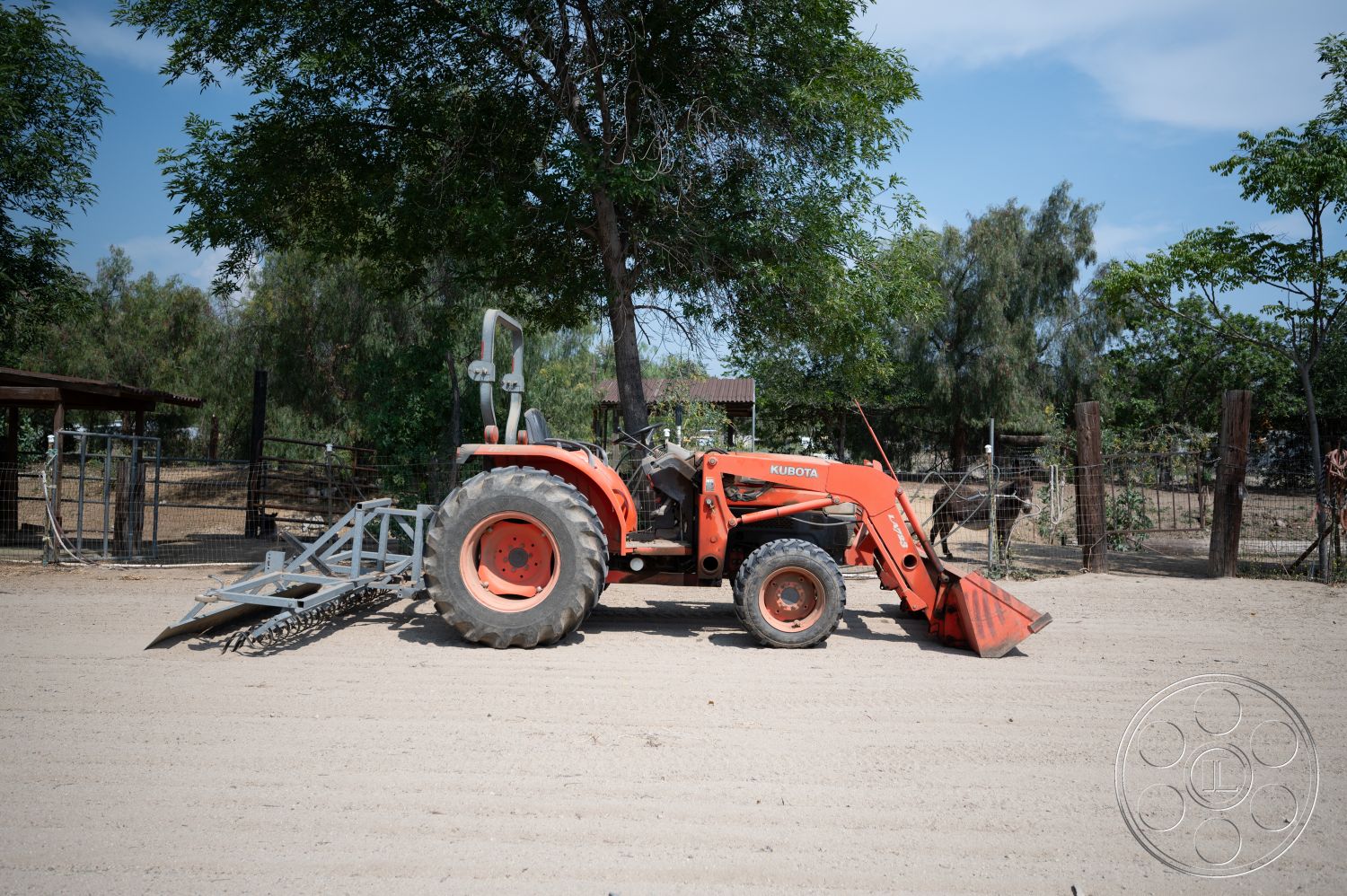 Ranch 6 - exterior, wooden fencing, surrounding green trees, rustic farm setting, dirt ground surface, orange agricultural tractor, Kubota tractor, metal plow attachment