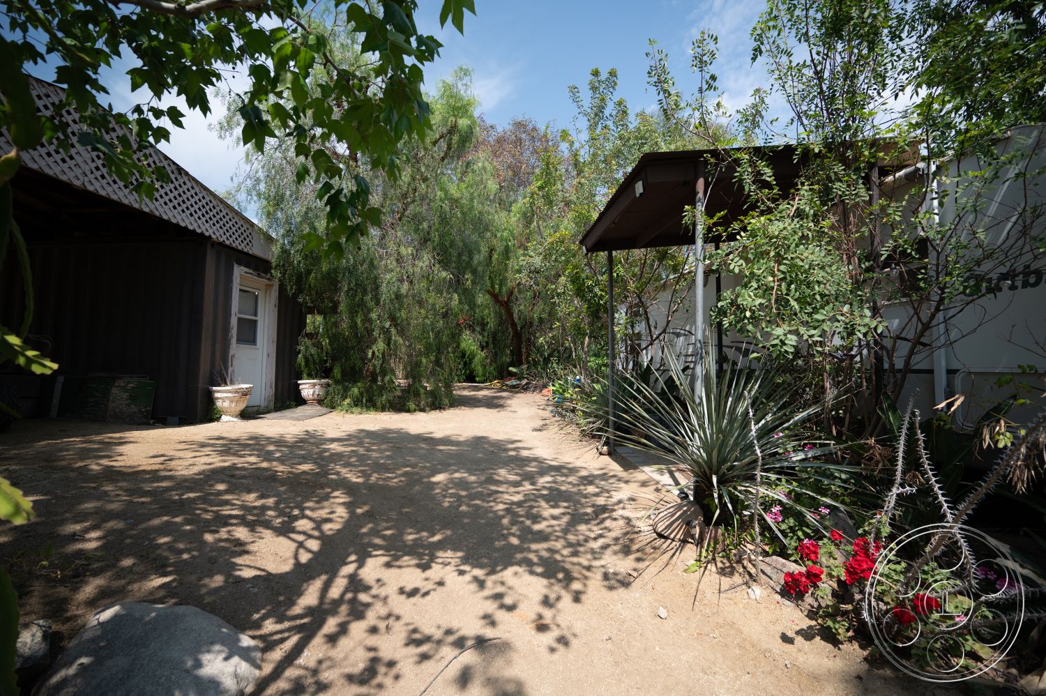 Ranch 6 - exterior, tropical plants, green shrubbery, flowering plants, gravel driveway, outdoor path, landscaped garden bed, wooden porch roof, painted metal siding, large ornamental planters, shade tree canopy, boulders in landscape