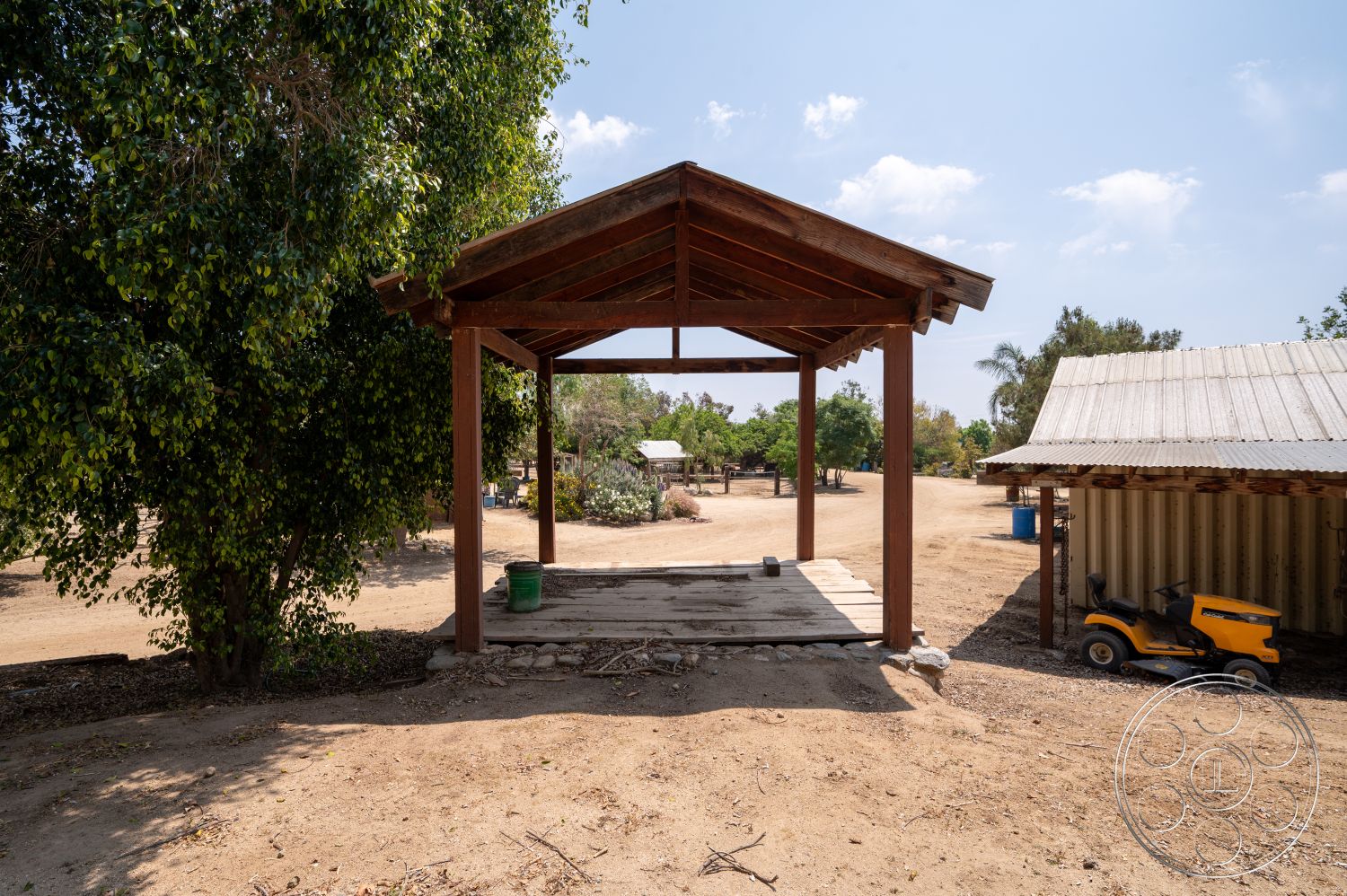 Ranch 6 - exterior, green foliage, open-air design, surrounded by trees, gravel ground, rustic wooden beams, natural landscape setting, wooden gazebo structure, simple gabled roof, open floor area, desert-like environment, nearby shed structure