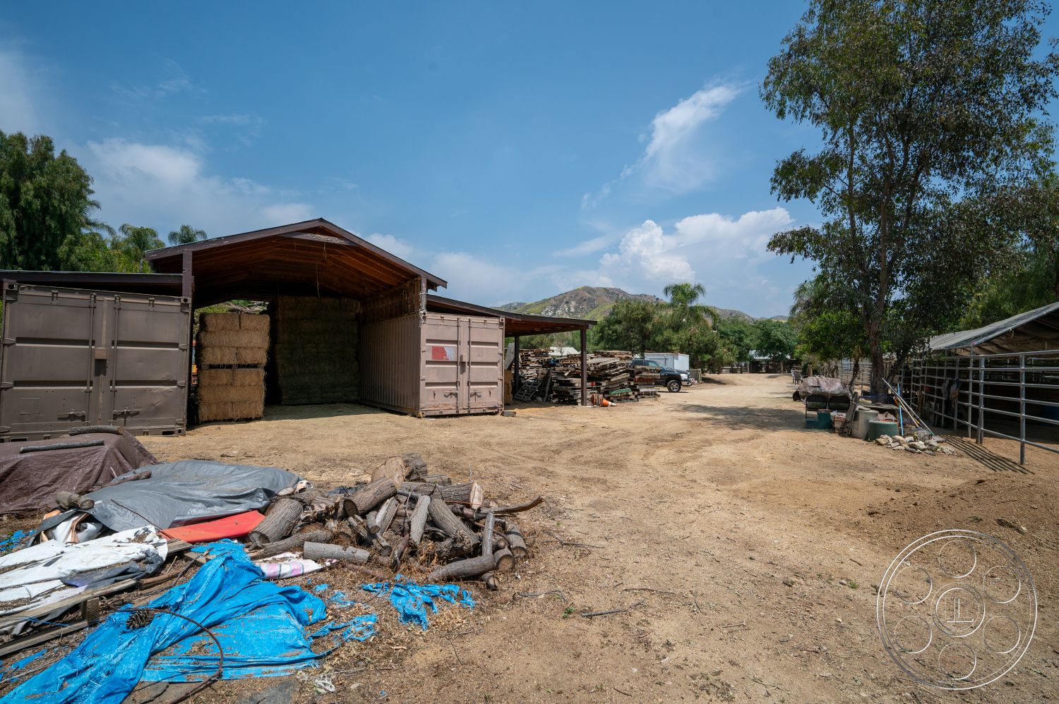 Ranch 6 - exterior, exposed wooden beams, mountain backdrop, rural setting, dirt driveway, hay bales, metal shipping containers, wood barn structure, stacked wood pile, rustic farm area