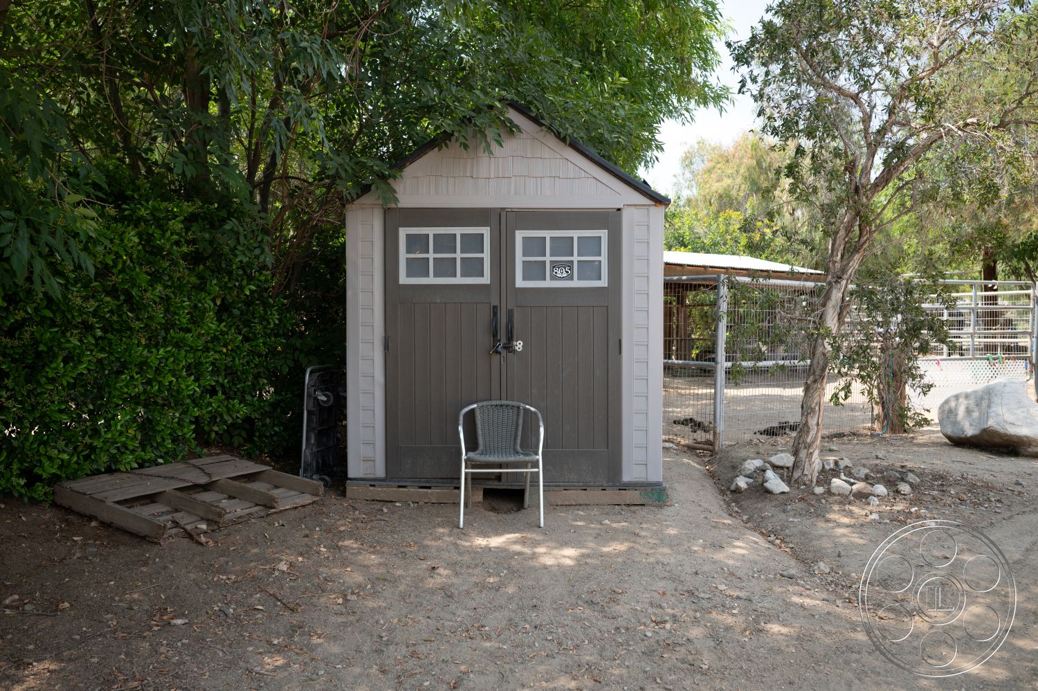 Ranch 6 - surrounding trees, wooden siding, double-hung windows, gable roof, gravel ground surface, exterior shed, gray painted doors, wooden pallet flooring, landscaped with shrubs, sitting area with chair