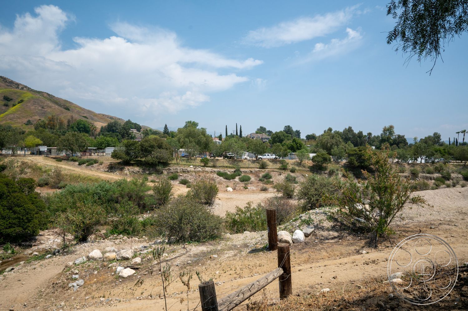 Ranch 6 - exterior, dirt path, natural vegetation, mountain backdrop, scenic landscape, rolling hills, lush trees, wooden fence posts, sparse shrubbery, blue sky with clouds