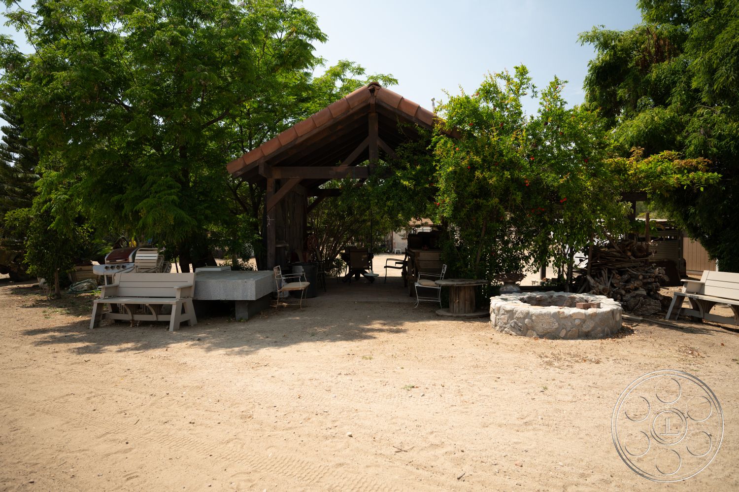 Ranch 6 - exposed wooden beams, terracotta tile roof, desert environment, concrete fire pit, rustic outdoor setting, wooden seating area, lush greenery landscaping, gravel outdoor flooring, covered outdoor space, wooden post pavilion