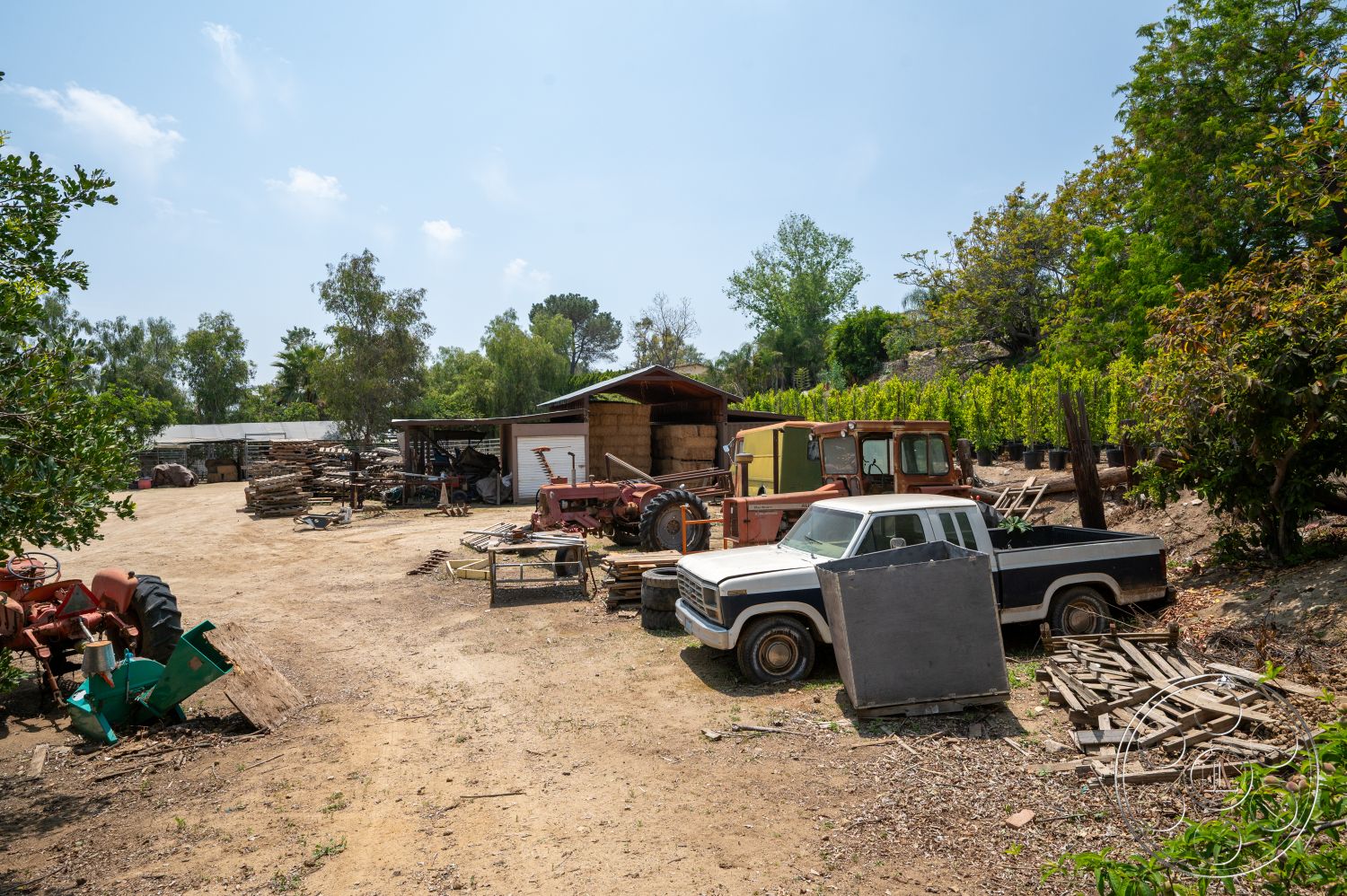 Ranch 6 - exterior, open outdoor space, gravel ground, dirt driveway, landscape trees, wooden storage shed, rustic tractor, white pickup truck, green farming equipment, stacked wooden pallets, farmyard setting, vineyard background, rusty machinery