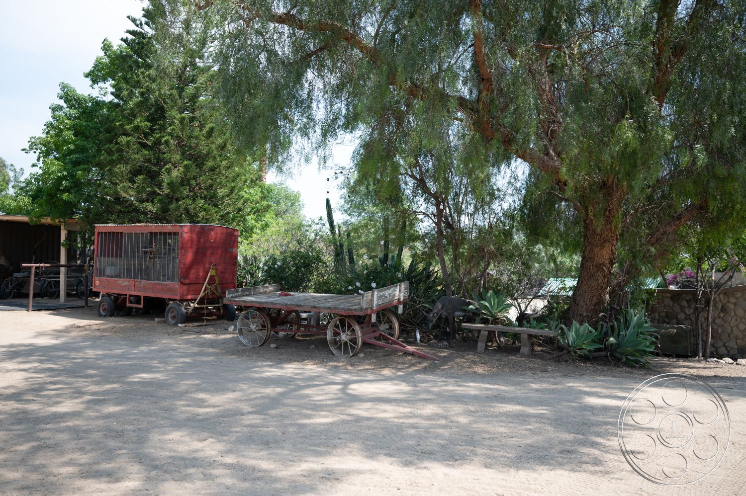 Ranch 6 - exterior, stone wall, surrounding greenery, dirt ground, rustic outdoor setting, wooden wagon, metal animal pen, large leafy trees