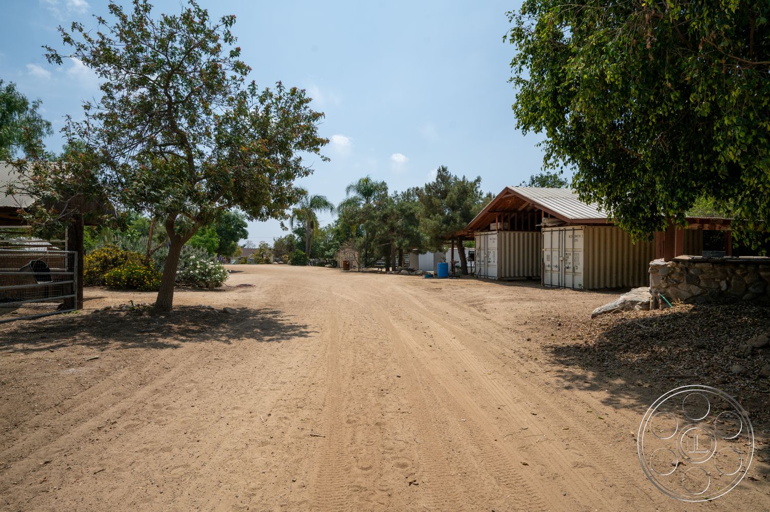 Ranch 6 - exterior, open-air environment, desert landscape, dirt driveway, tree shade, landscaped garden area, rural farmhouse setting, metal storage containers, wooden structure with metal roof