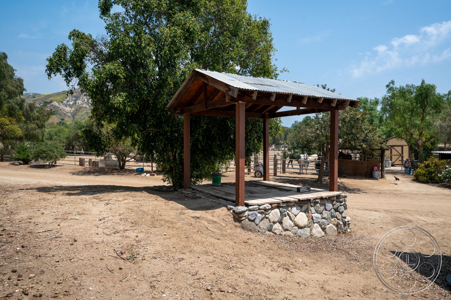 Ranch 6 - exterior, surrounding trees, natural stone wall, stone base, wooden pergola structure, metal corrugated roof, dirt ground surface, landscape hillside, open farm environment