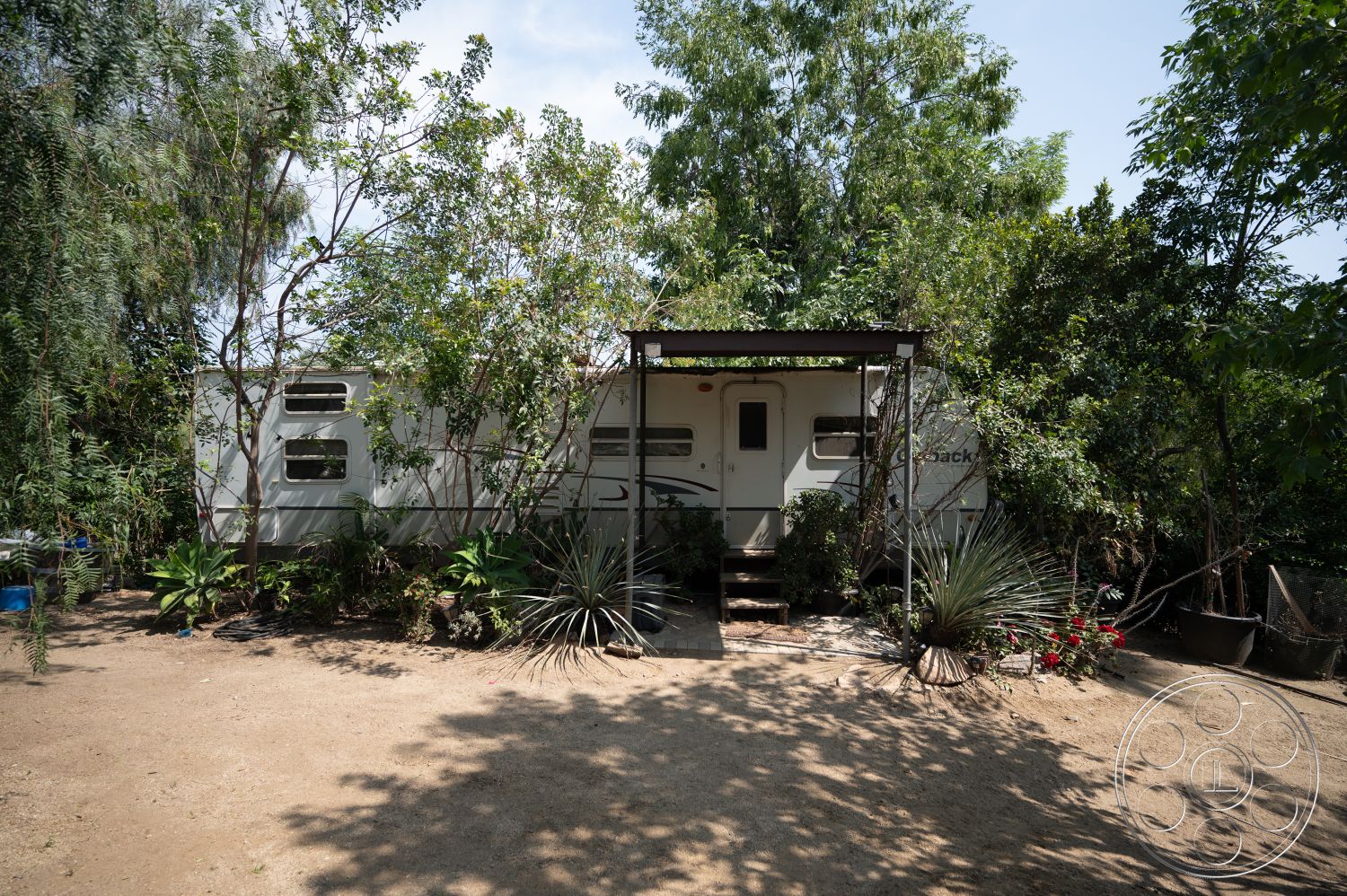 Ranch 6 - exterior, potted plants, surrounding greenery, wooden steps, front porch, mobile home, rustic outdoor setting, sunny outdoor environment, landscape vegetation, aluminum siding, bare dirt yard