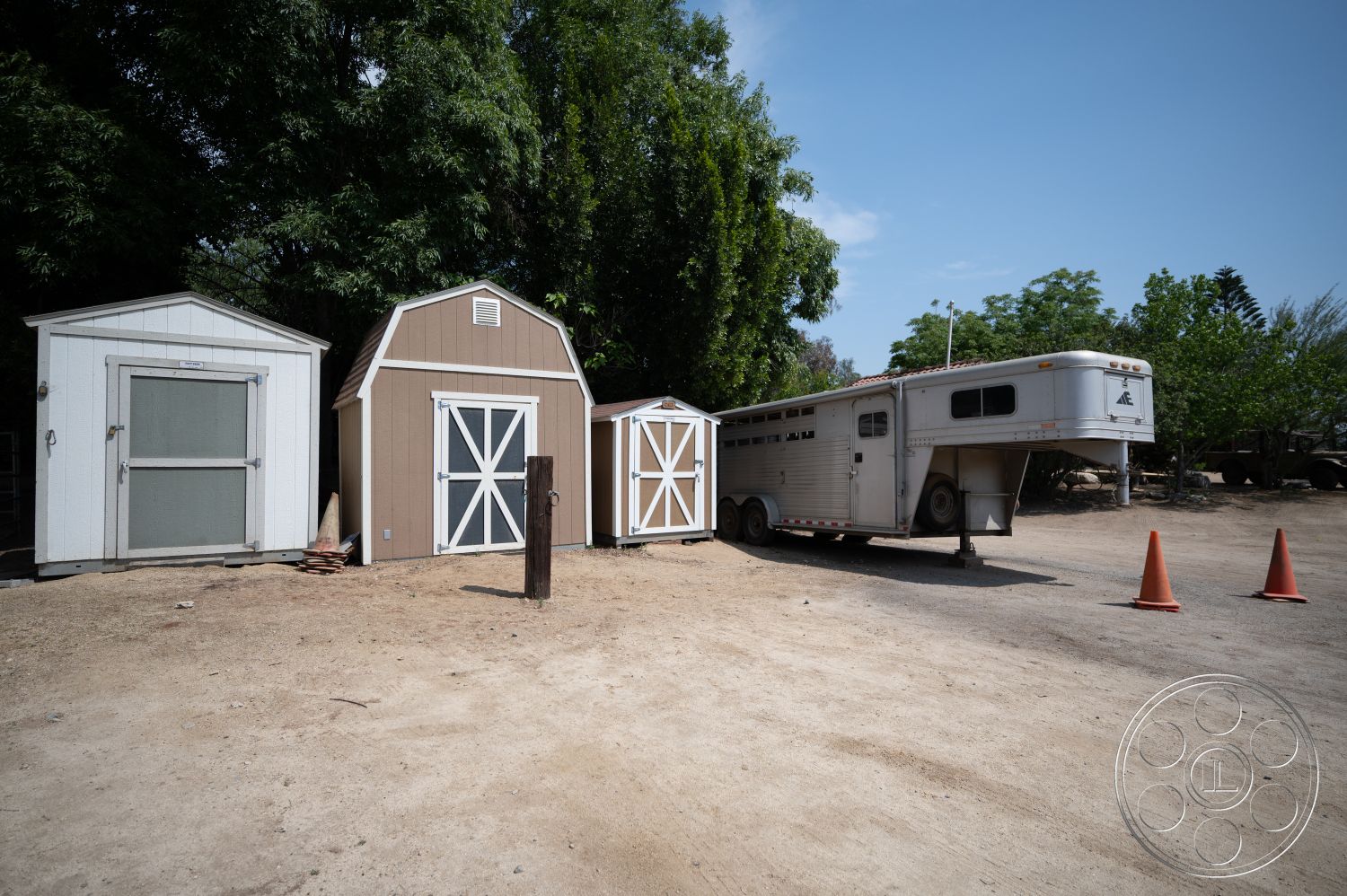 Ranch 6 - rural setting, orange traffic cones, outdoor environment, wooden shed exterior, brown wood barn-style shed, white wooden shed with glass door, aluminum horse trailer, dirt driveway surface, green foliage surrounding, concrete support post