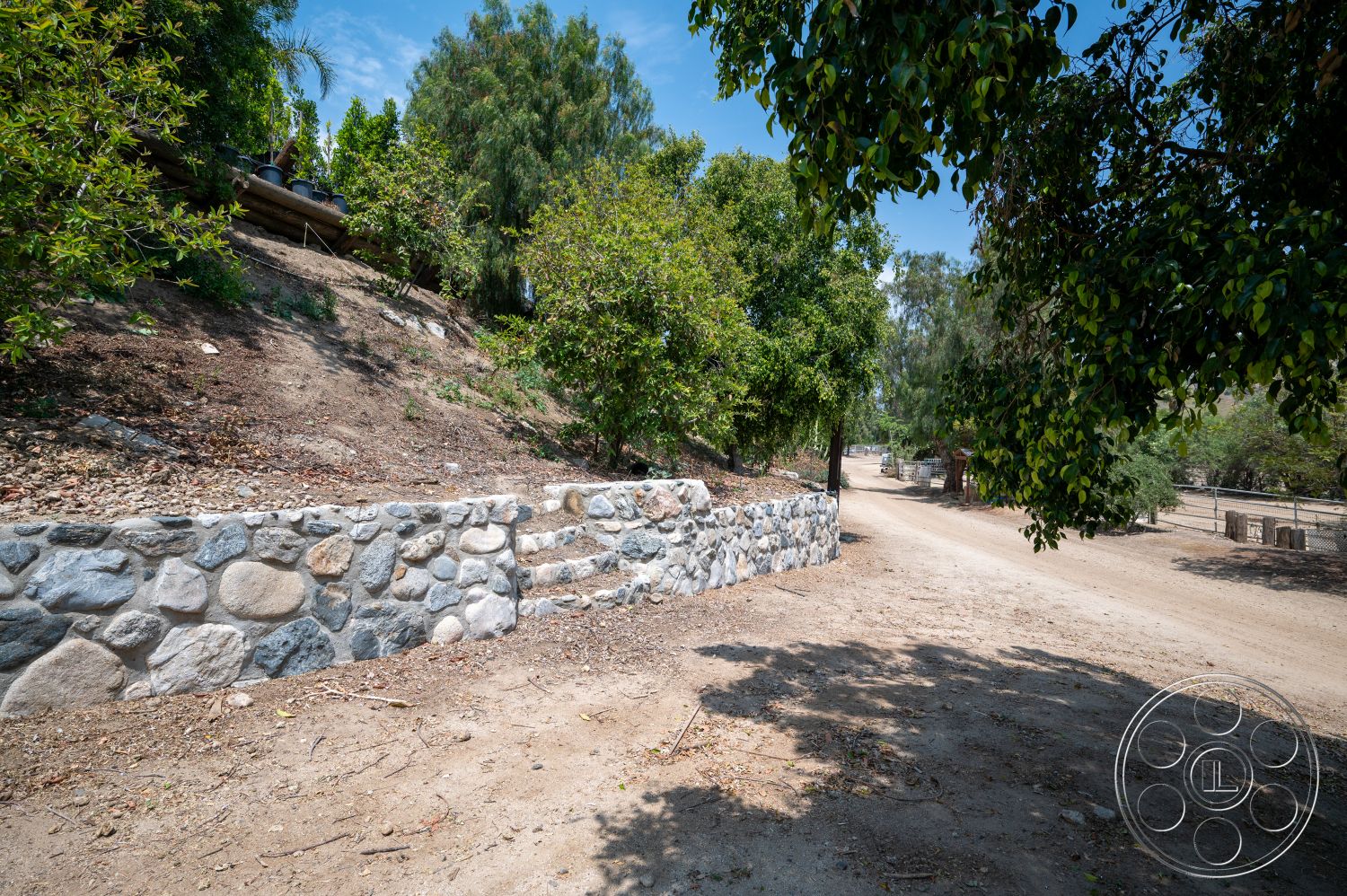 Ranch 6 - exterior, sloped terrain, dirt road, mountainous background, natural stone retaining wall, sunny day setting, rounded stone wall, landscape with dirt pathway, vegetation with trees