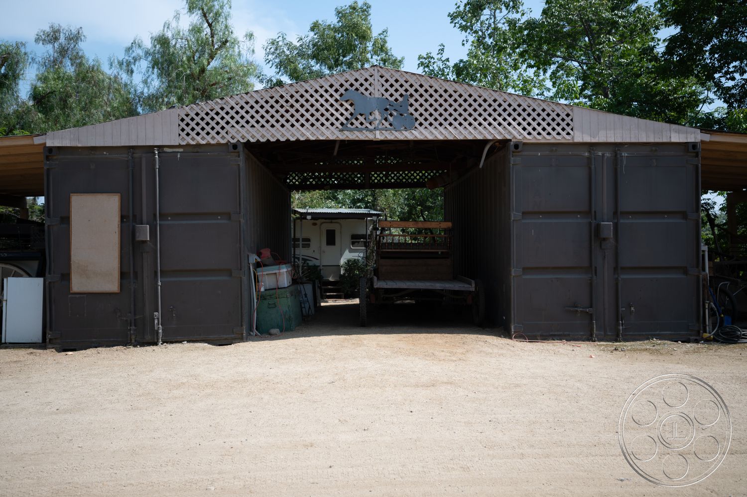 Ranch 6 - exterior, wooden beams, dirt driveway, surrounding vegetation, agricultural setting, open garage entrance, metal container structure, wood lattice roof, dark metal siding, large storage area