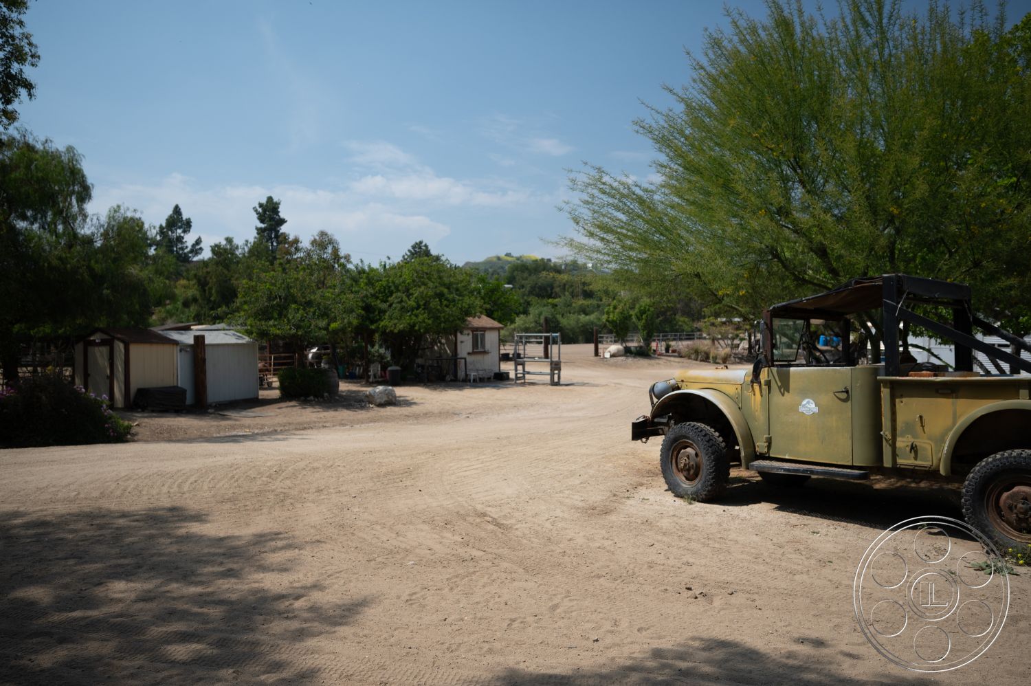 Ranch 6 - clear blue sky, green trees, dirt driveway, minimal landscaping, sunlit environment, rural farmhouse setting, wooden storage shed, covered outdoor area, dusty terrain, vintage pickup truck