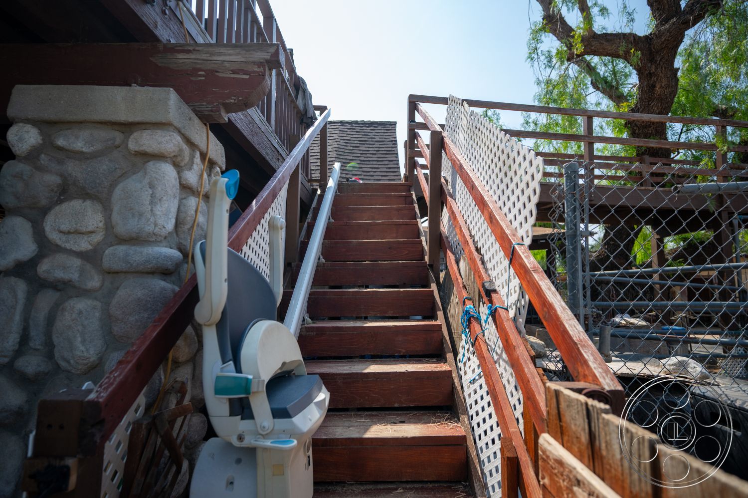 Ranch 6 - exterior, textured stone wall, wooden stairs, wooden handrails, chain-link fencing, stone support columns, painted wooden deck, metal stair lift, wood lattice panels, natural tree shade