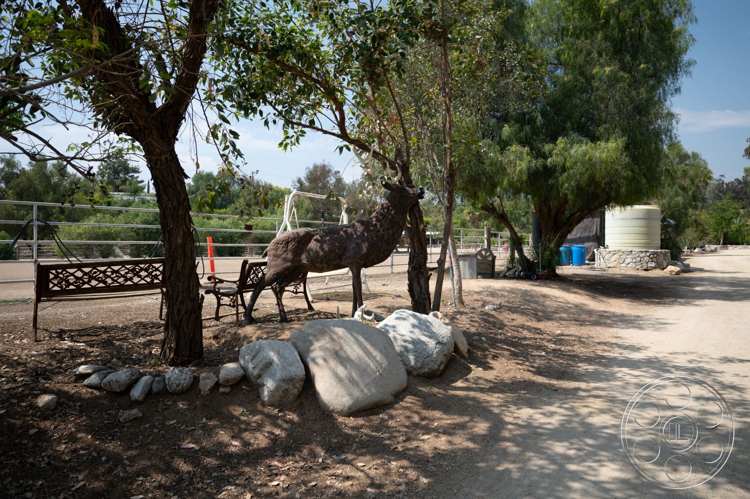 Ranch 6 - exterior, green foliage, metal fence, metal bench, dirt pathway, wooden tree trunk, natural stone landscape rocks, decorative deer statue, water storage tank, stone foundation wall